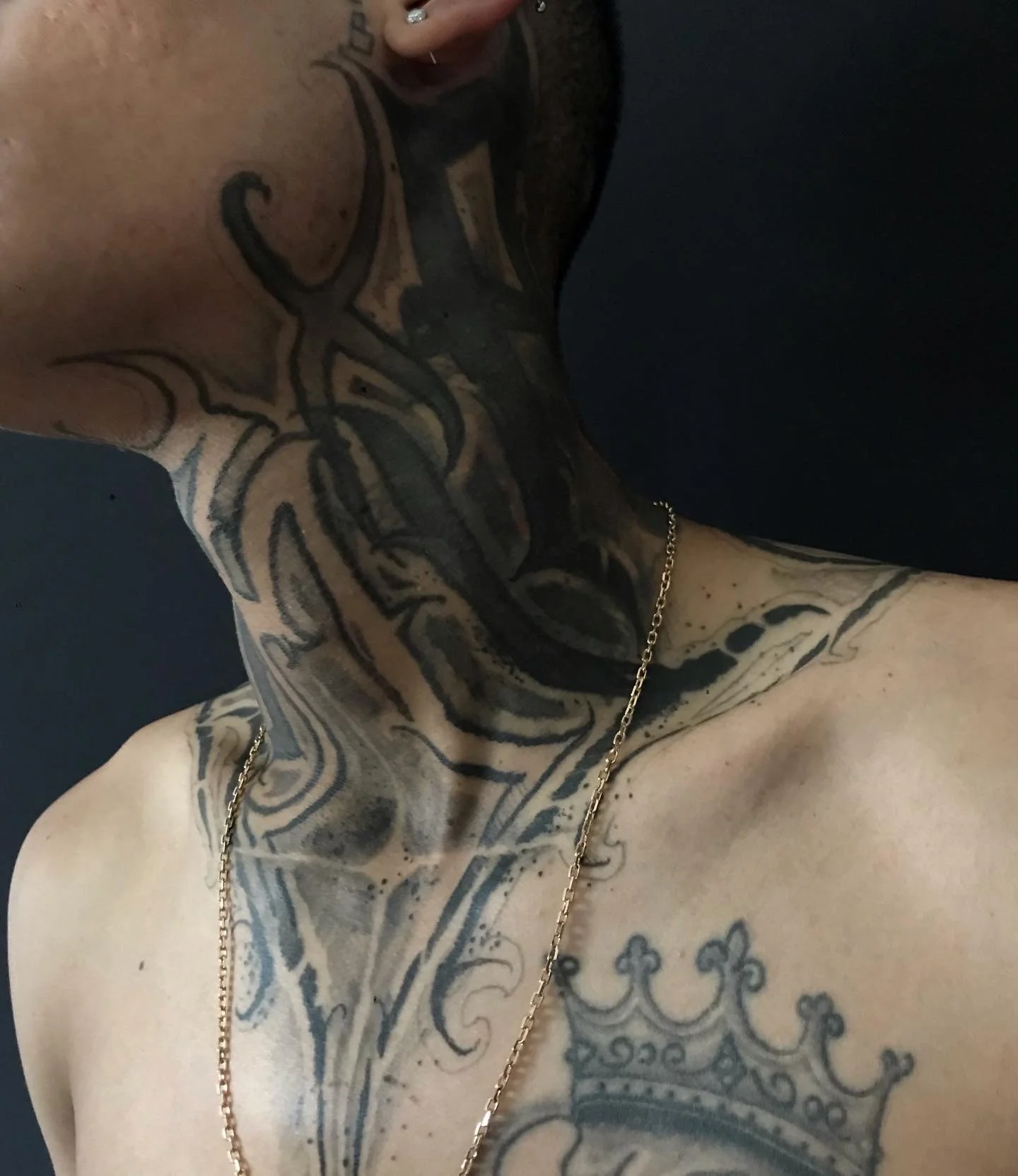 Close-up of a person's chest, neck, and part of the face showing tattoos, gold chain necklace, and earrings against a dark background.