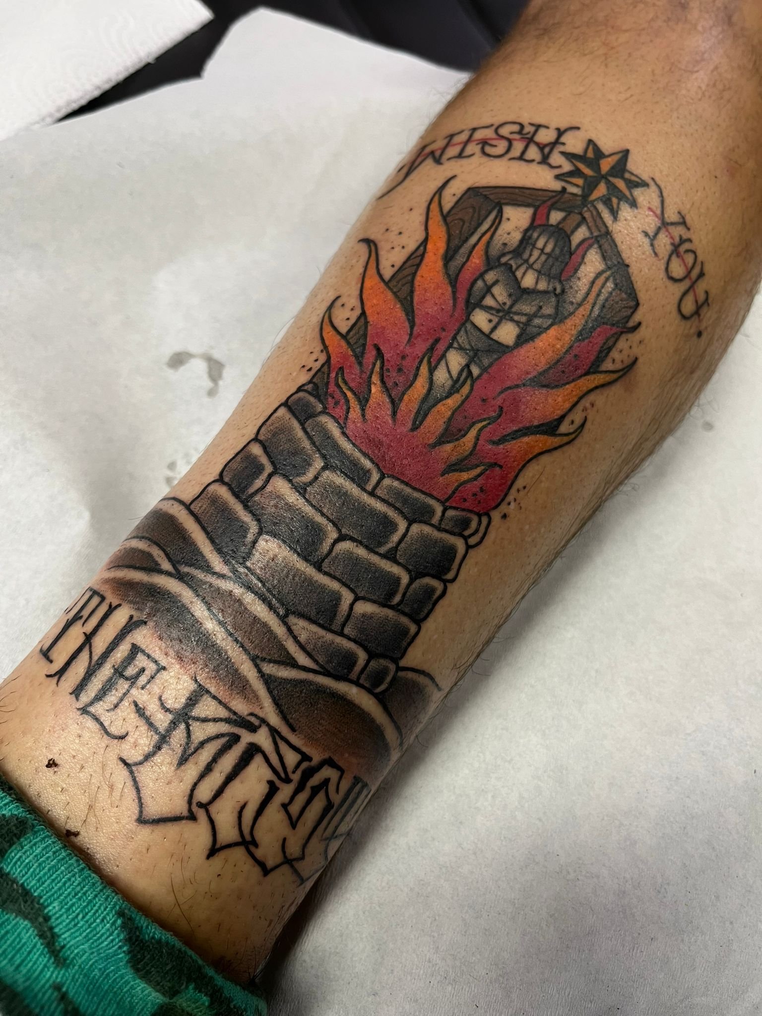 A tattoo of a torch with flames on a person's forearm. The torch is decorated with brick-like patterns and the flames are red and orange. Text above and below the torch reads 'Flesh & Bone' and 'You'. There is a star design near the top.