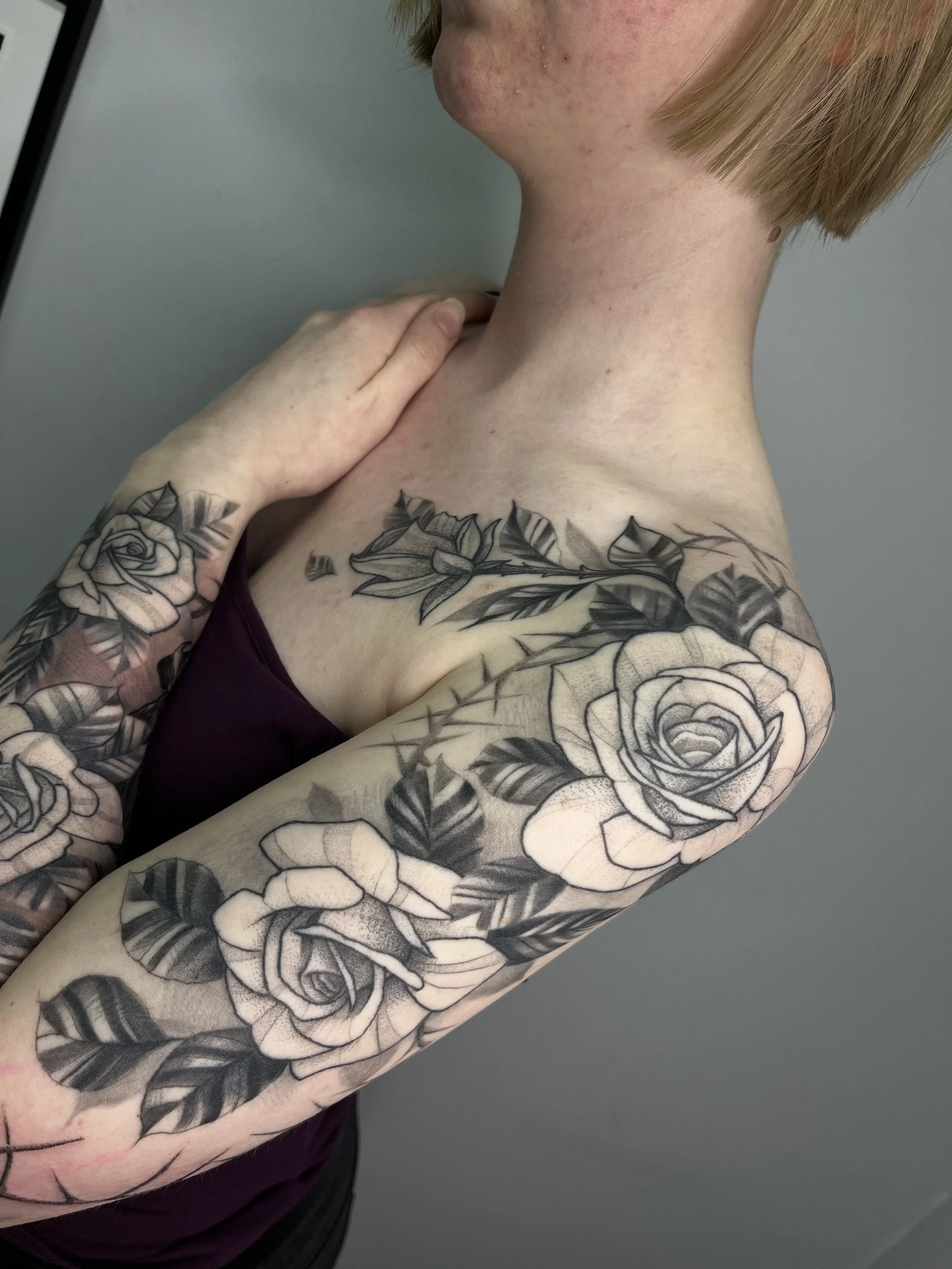 A person with short hair and tattoos on their arm and shoulder, wearing a purple top, touching their neck and collarbone area.