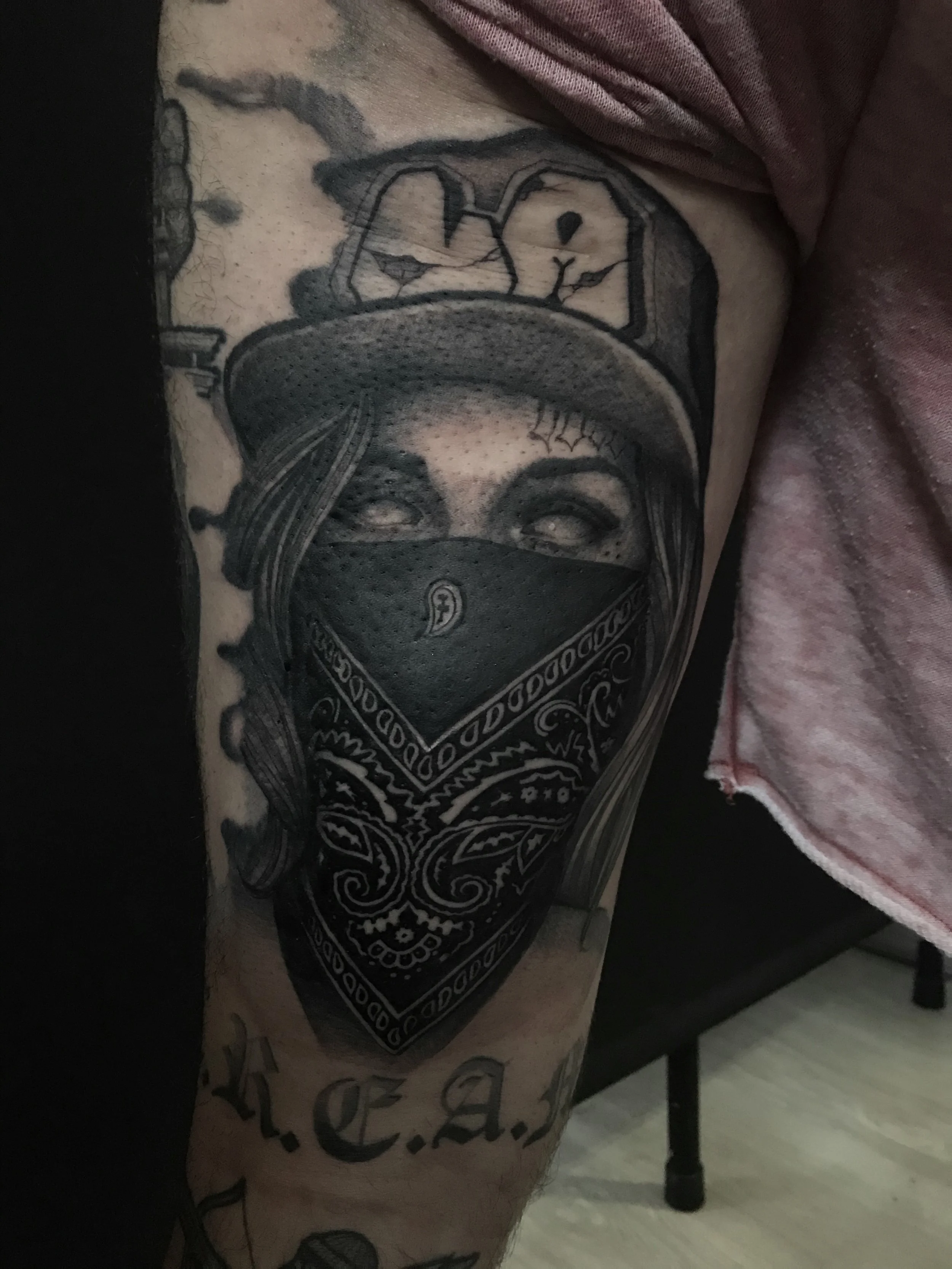 Tattoo of a person wearing a bandana covering their nose and mouth, with a sombrero and a cartoon face with smiling eyes and an open mouth underneath, on a person's arm.