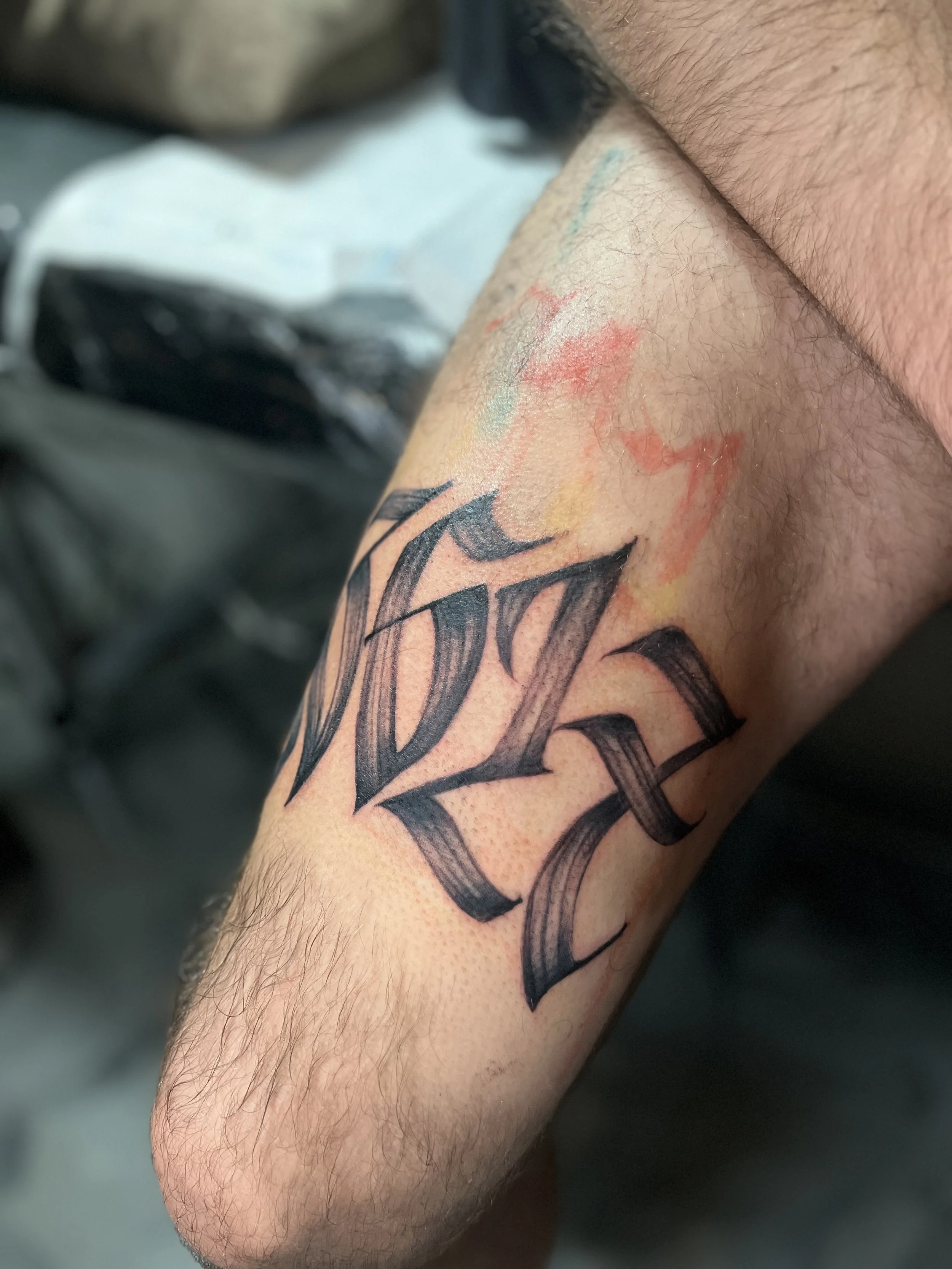 Close-up of a tattoo on a person's forearm with black Gothic-style lettering, some redness and watercolor-like ink smudges around it.