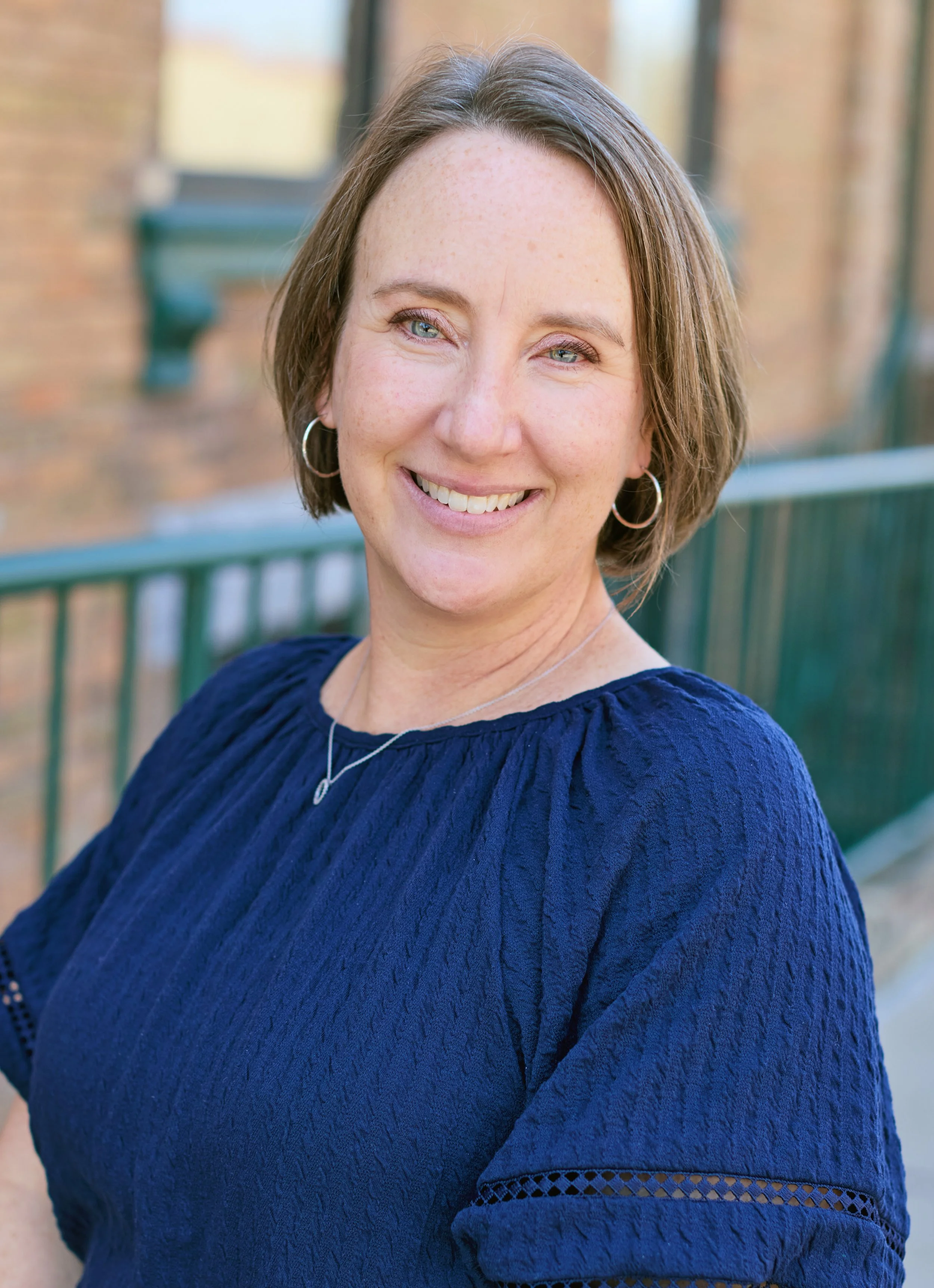A woman with short brown hair and blue eyes smiling outdoors, wearing a navy blue textured top, hoop earrings, and a necklace, with a green railing and brick building background.