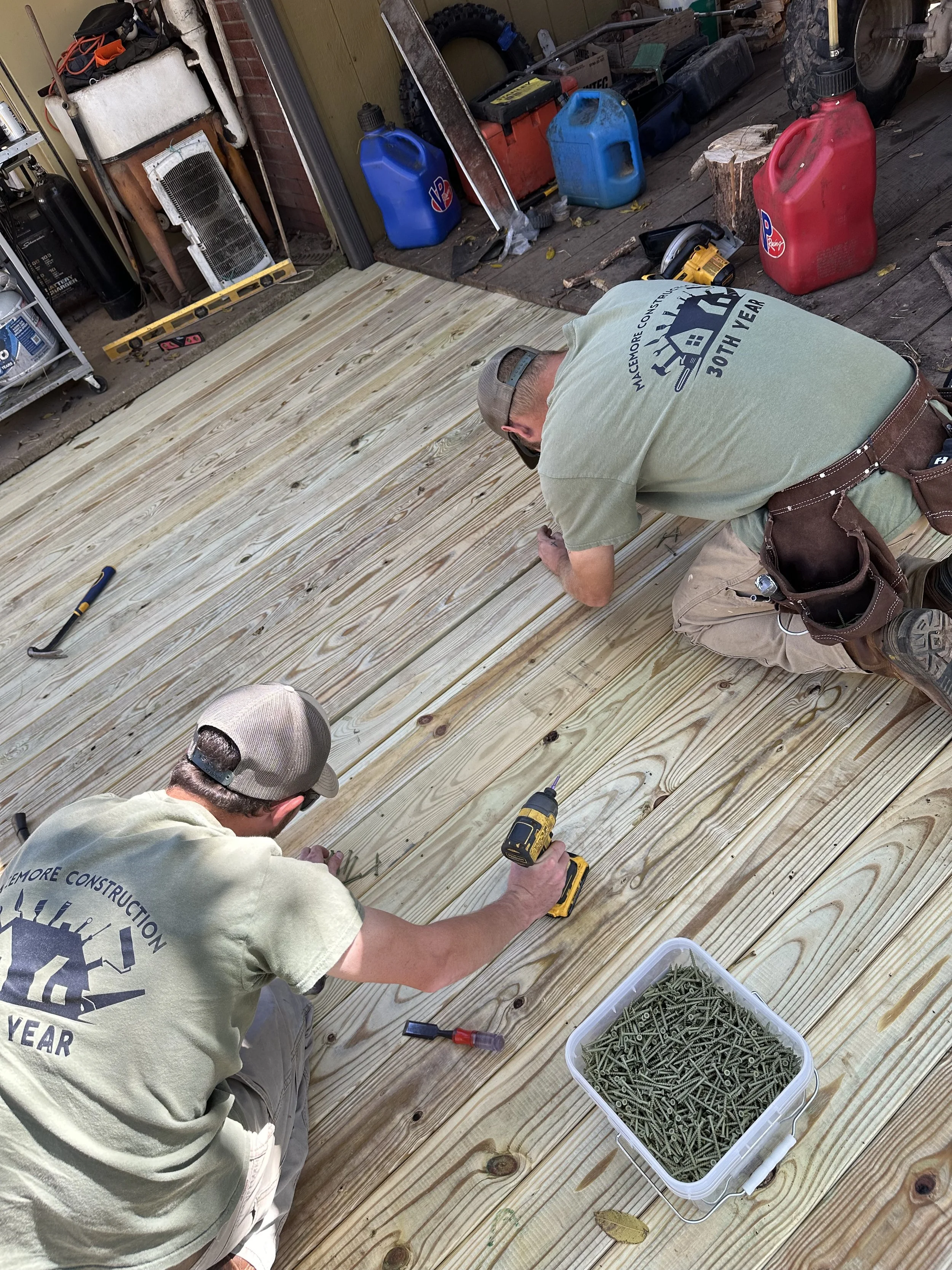 Two men working on building a wooden deck, using tools and screws, with construction supplies and fuel containers in the background.