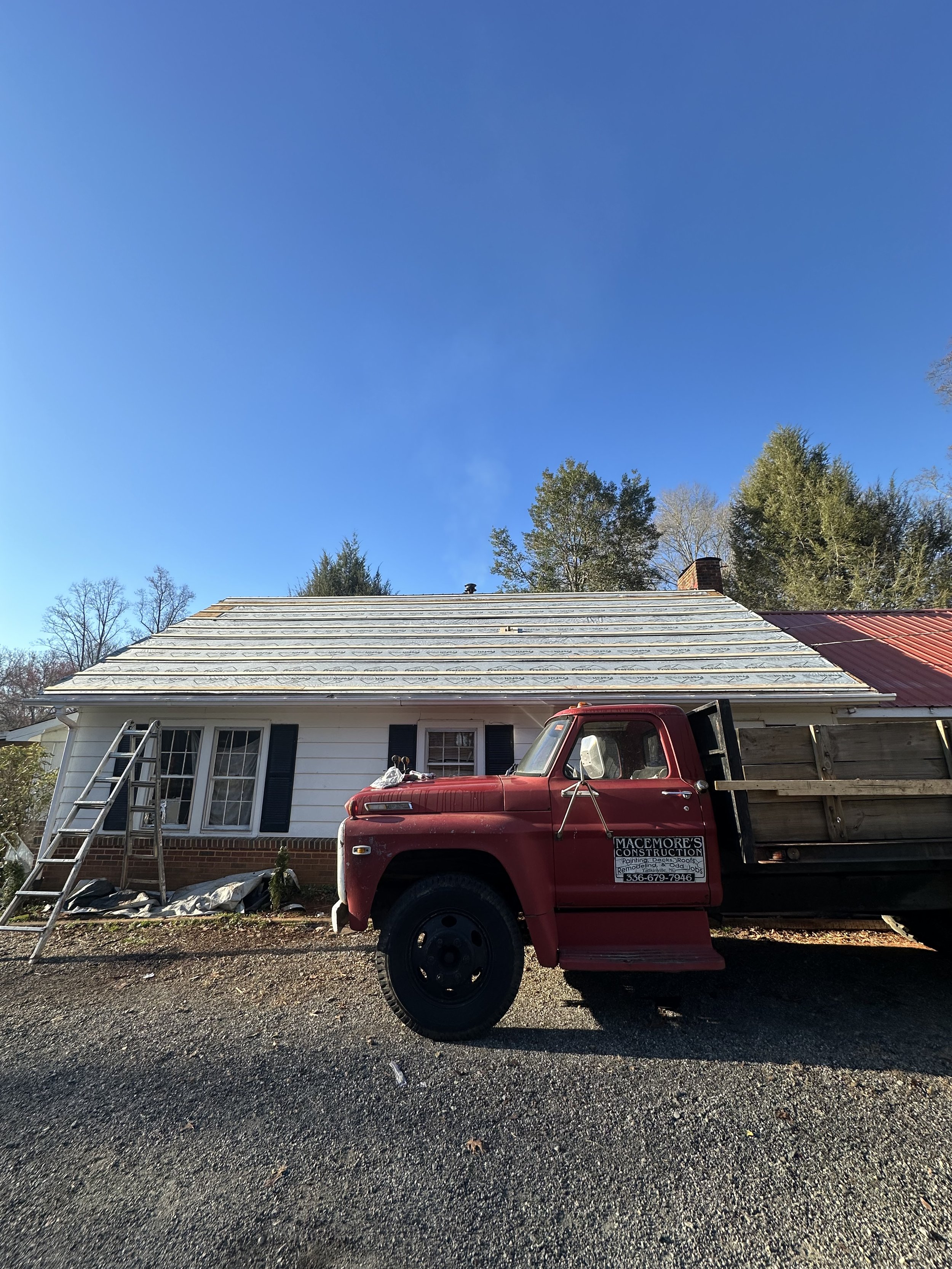 A house with a white exterior, black shutters, and a brick foundation, under repair with a new roof being installed. A red flatbed truck is parked in front of the house, with a ladder leaning against the left side, and construction materials and tool