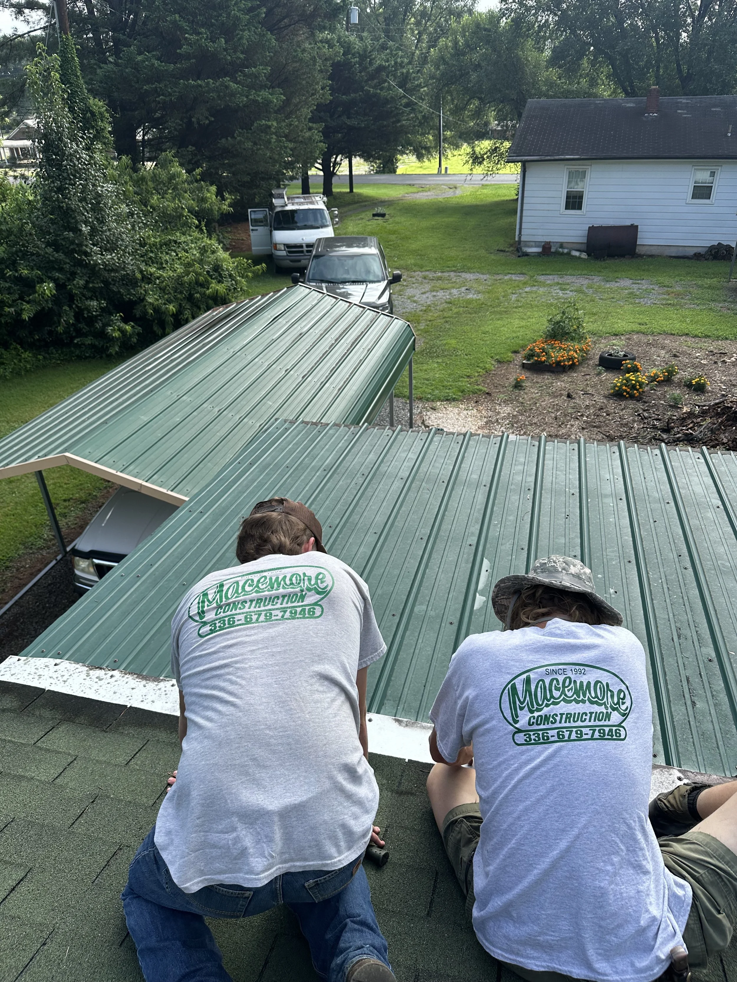 Two workers from Macemore Construction installing a green metal roof on a building, with one person wearing a tan hat and both in gray shirts with company logo, on a residential rooftop.