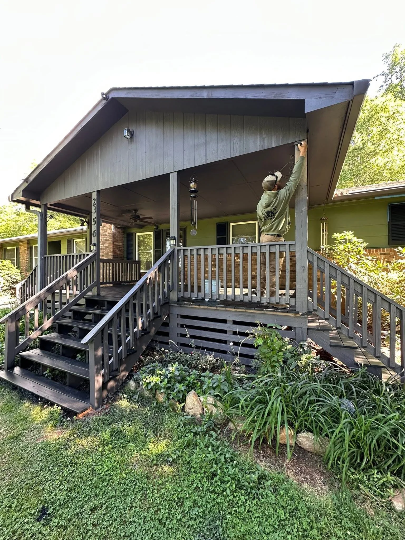 A man painting the upper part of a house’s exterior wall on a covered porch with dark green vertical siding, while standing on a wooden deck accessed by stairs, surrounded by greenery and garden plants.
