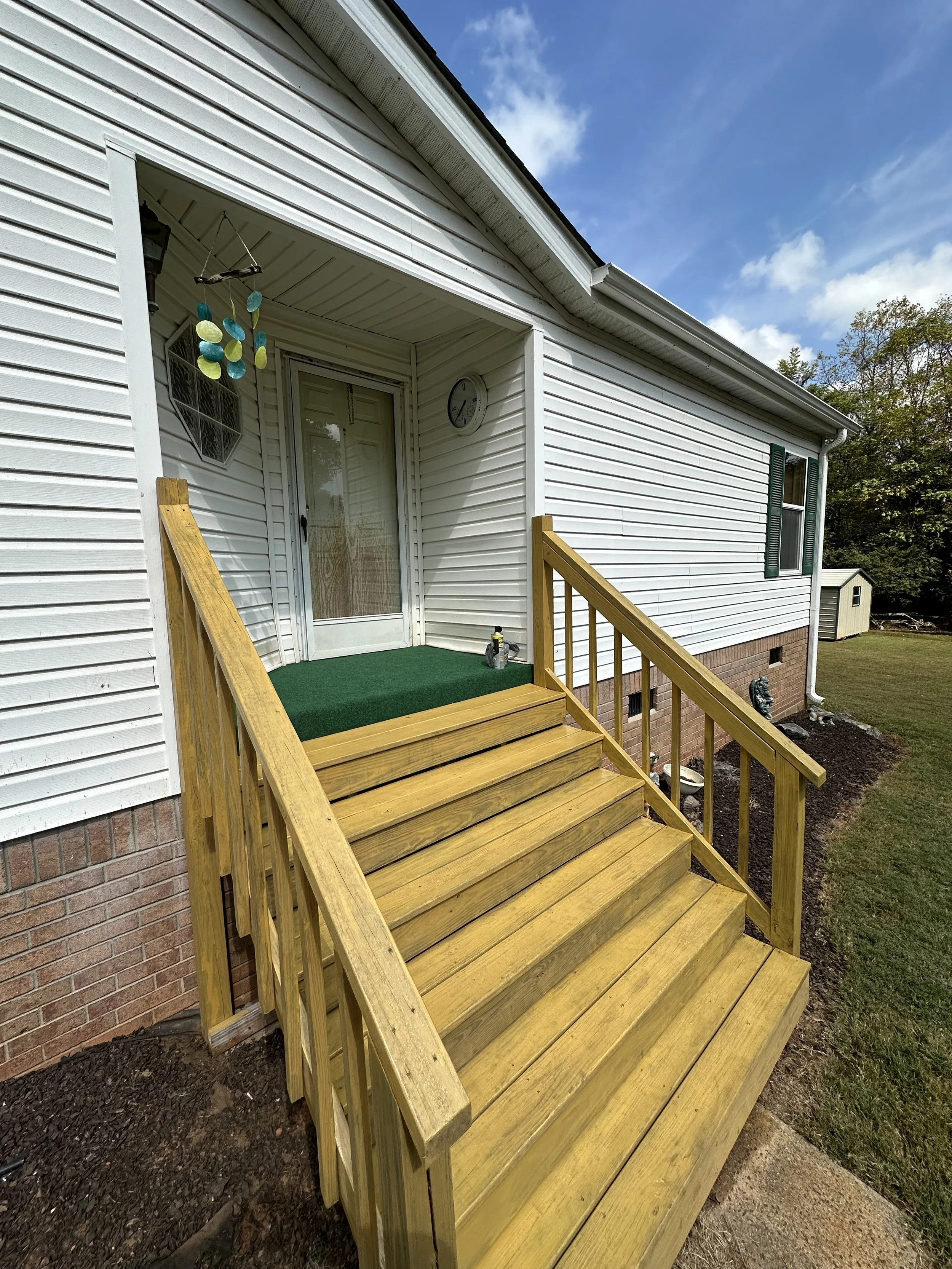 Exterior view of a front porch with wooden stairs, white siding house, and a small green welcome mat, under a partly cloudy sky.