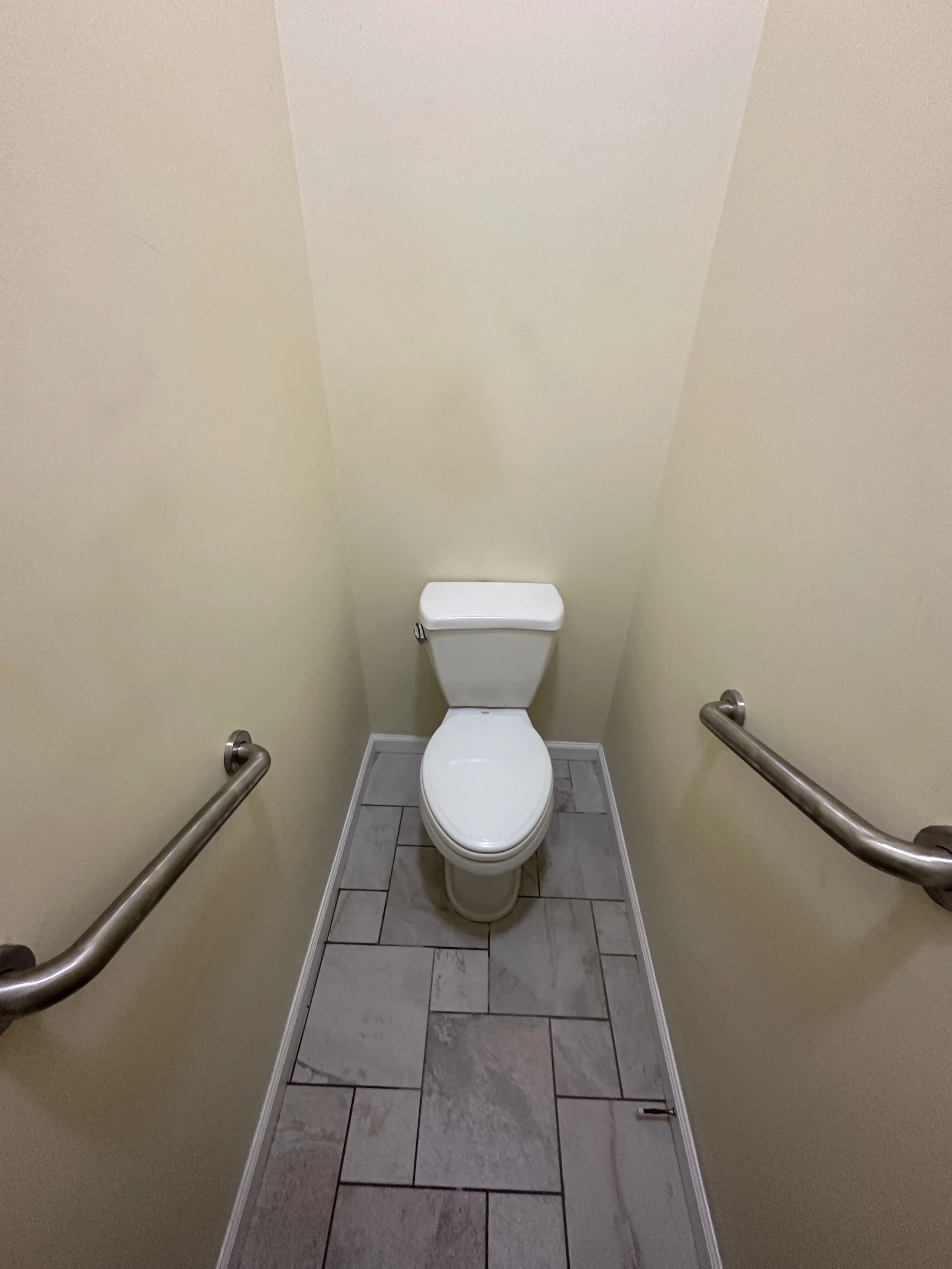A small, narrow bathroom with a white toilet, beige walls, and gray tile flooring. Metal grab bars are mounted on both sides of the walls.