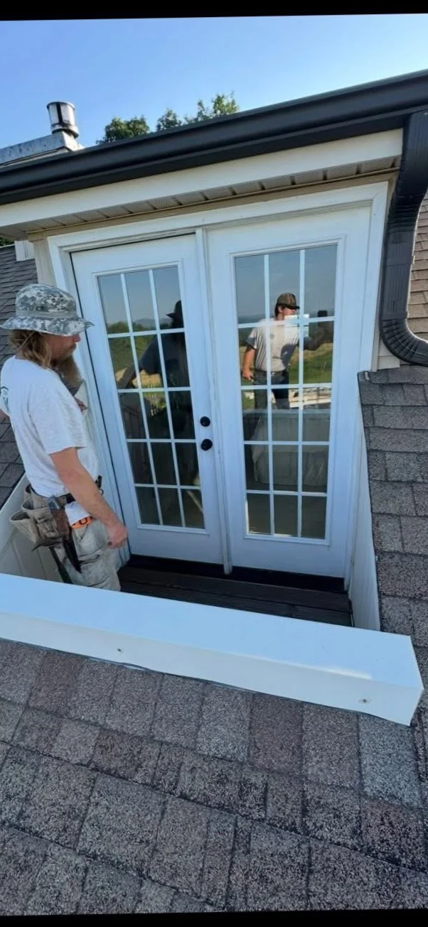 Person with a camouflage hat and work gear standing near glass double doors on the roof of a house, with another person visible through the doors reflected in the glass.