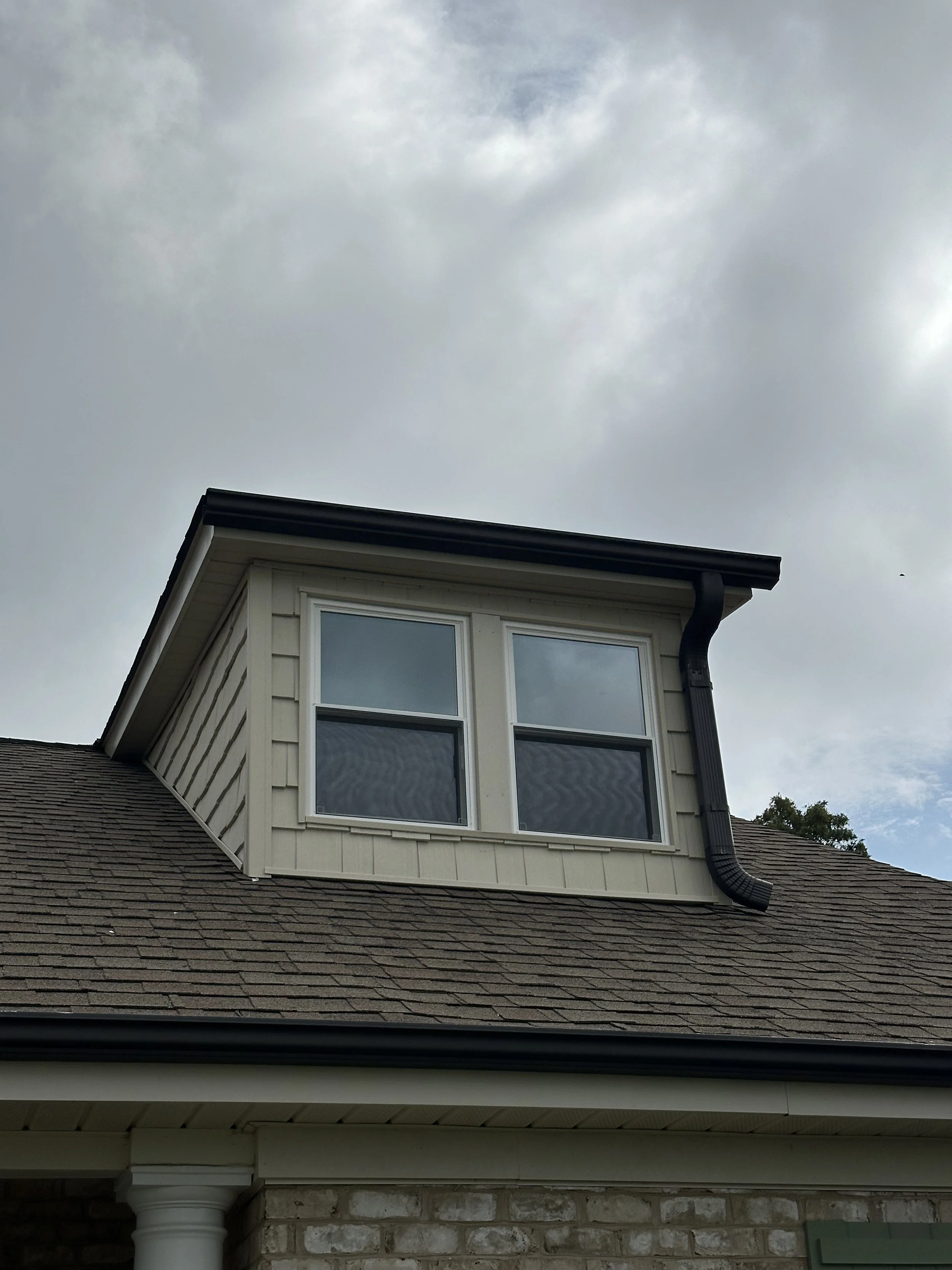 Close-up of a house's attic window with white trim, set under a gray, overcast sky, with a black gutter running along the roof.