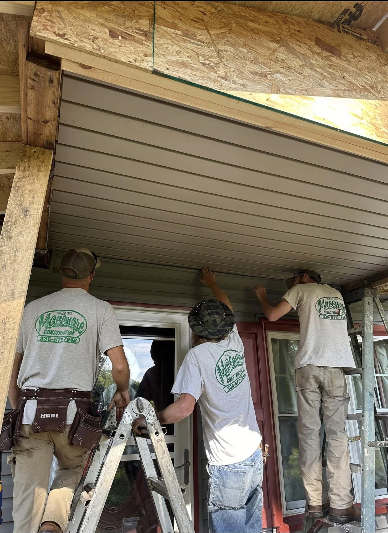 Three workers installing or repairing a porch ceiling on a house. One person on a ladder to the right, another in the middle reaching up to the ceiling, and the third on a stepladder, all wearing shirts with the logo 'Maccune Construction'.
