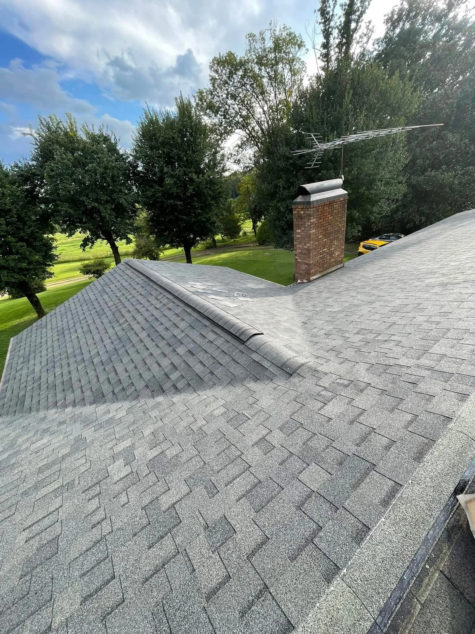 View of a house roof with gray shingles, a brick chimney, and a TV antenna, surrounded by green trees under a partly cloudy sky.