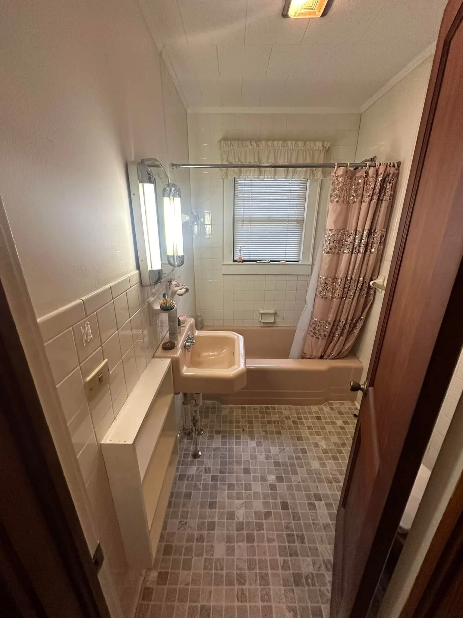 A small bathroom with a beige bathtub, a peach-colored sink, a window with blinds, a shower curtain with a sequin design, and a tiled floor.
