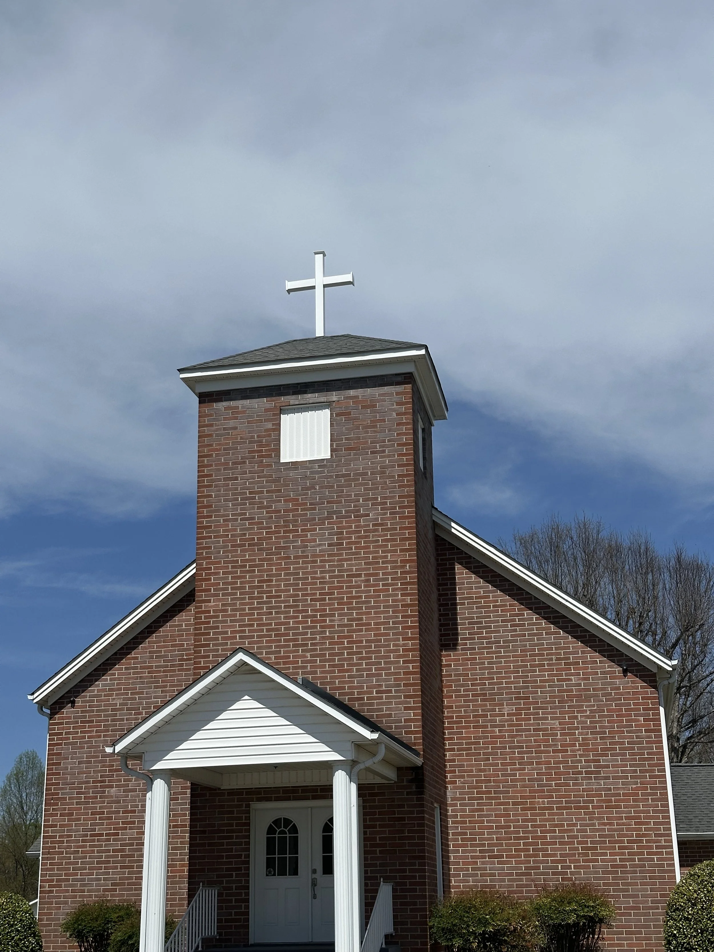 A brick church building with a white entrance door, white columns, and a small white triangular roof over the door. A tall brick steeple with a white cross on top is visible against a partly cloudy sky. Bushes are in front of the church.
