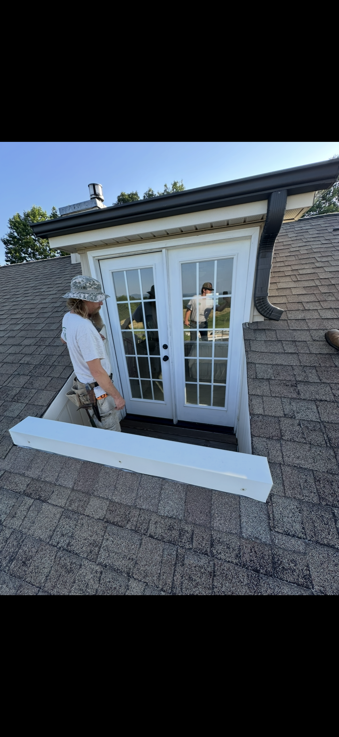 Two men inspecting a set of white French doors leading to a rooftop deck, with one man wearing a hat and proper work tools, on a shingled roof with a view of trees and blue sky.