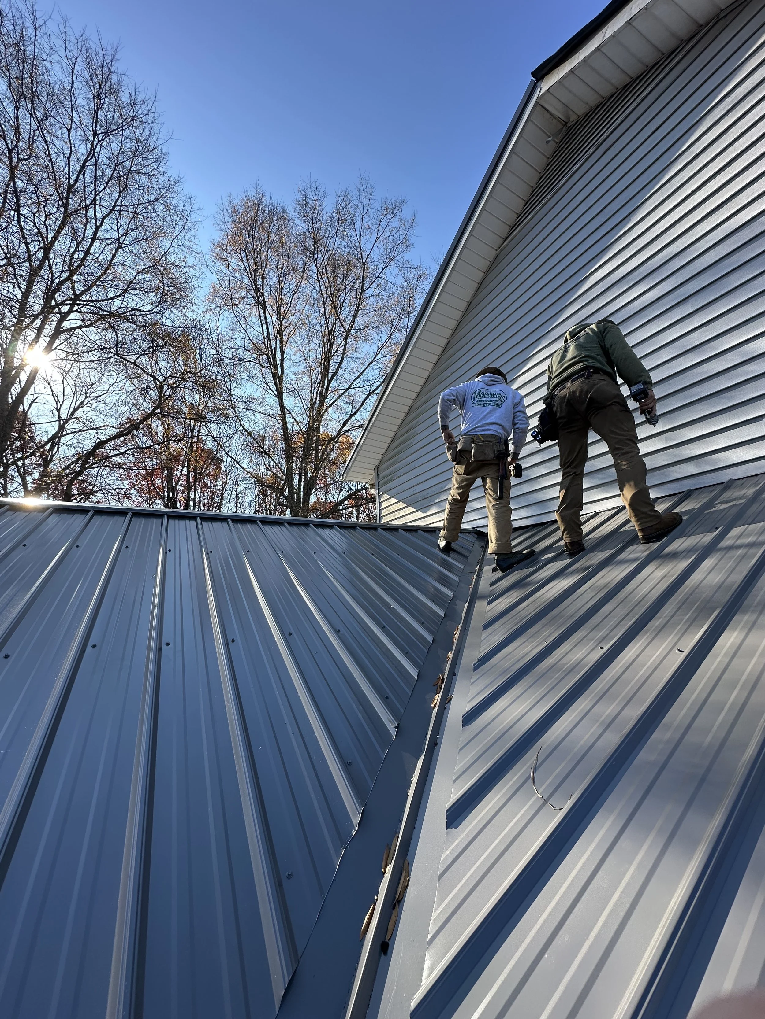 Two workers on a metal roof at a house, with leafless trees and a clear blue sky in the background.