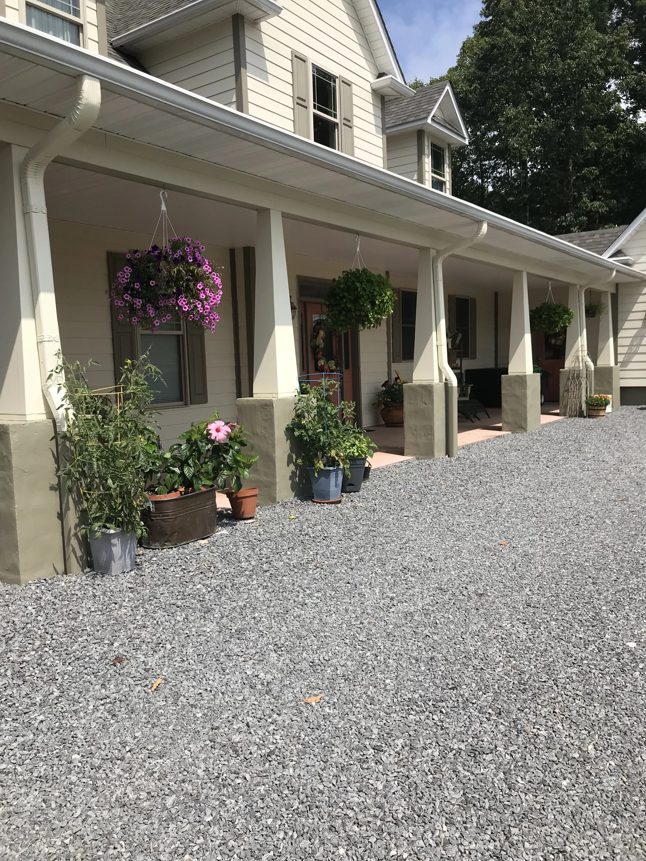 Front porch of a house with potted plants, hanging flower baskets, and gravel driveway.