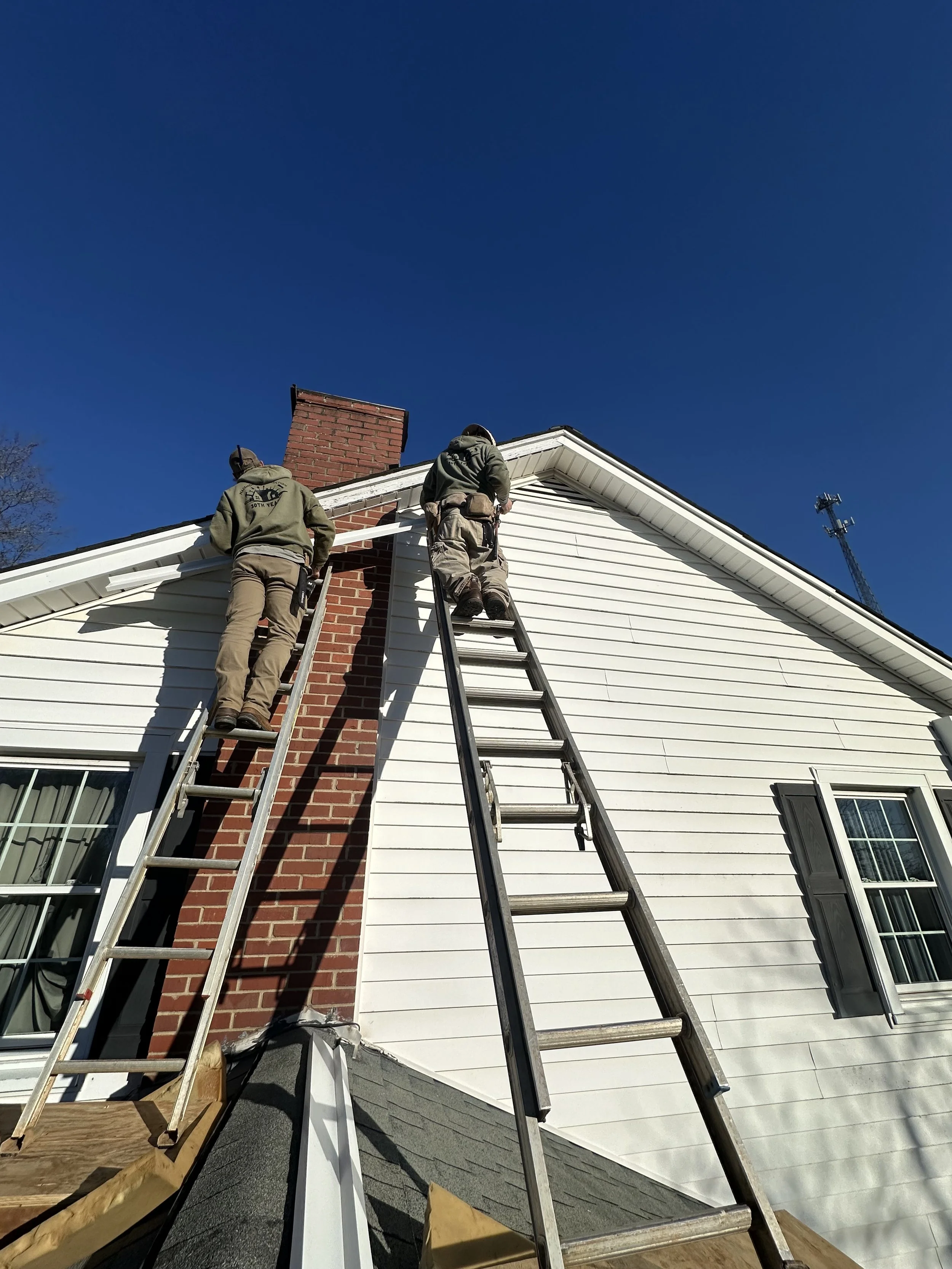 Two workers on ladders repairing the exterior of a house, standing near the roof and chimney under a clear blue sky.