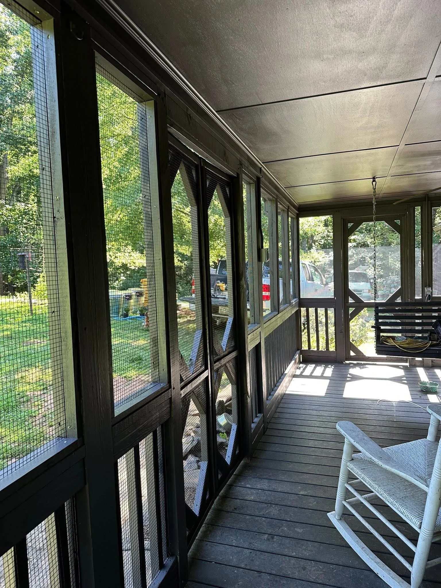 A screened porch with wooden flooring, black framing, a white rocking chair, parked cars outside, and a swing hanging from the ceiling.