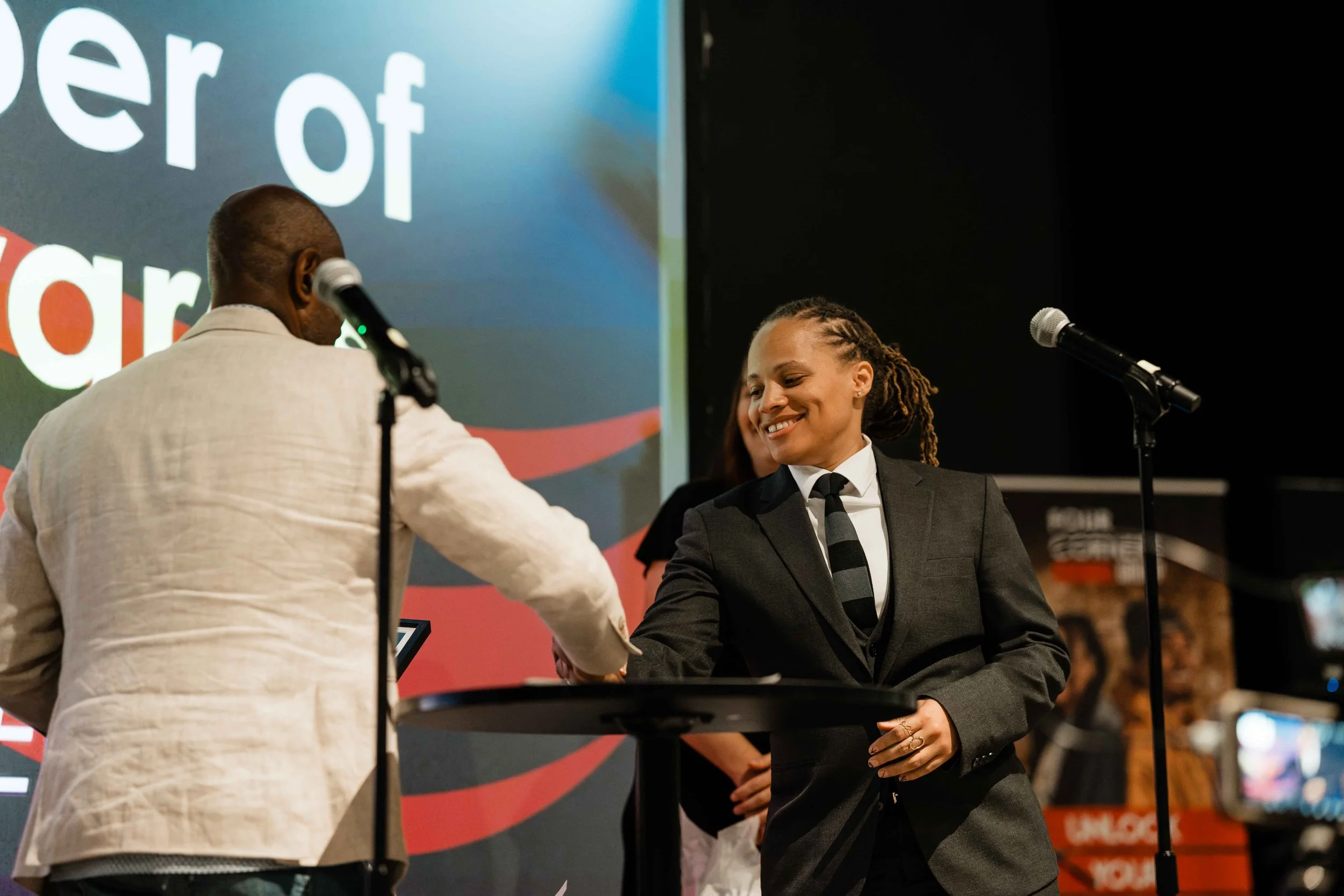 A woman in a black suit and striped tie smiling as she shakes hands with a man in a white jacket at a formal event, with microphones and a large screen in the background.