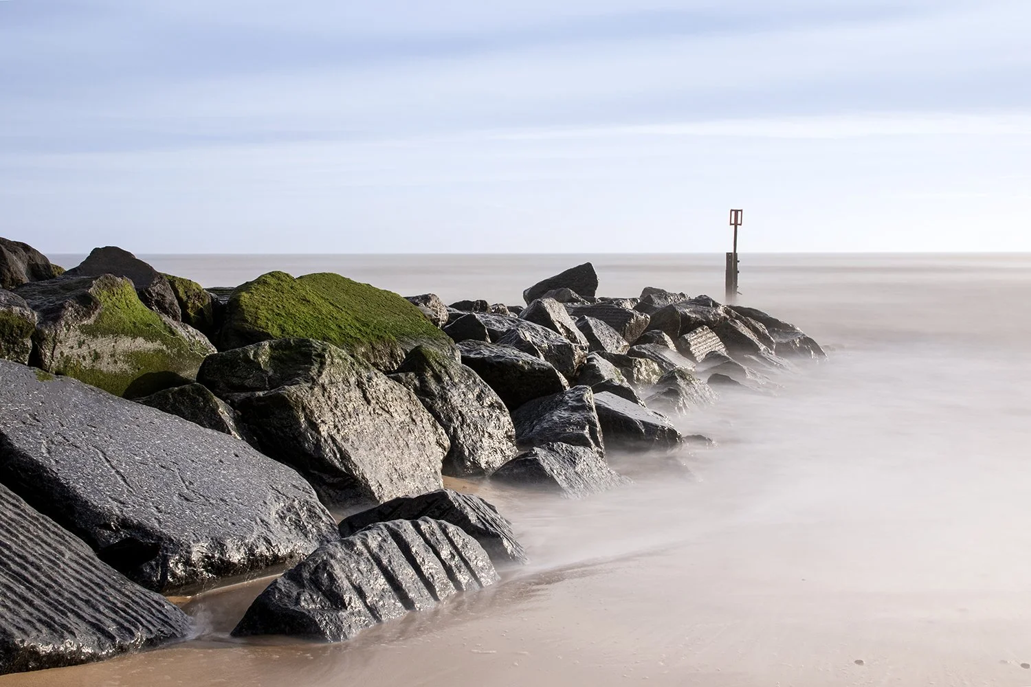 A rocky shoreline with moss-covered rocks leading into the ocean. Calm water and a cloudy sky with a navigational marker in the distance.