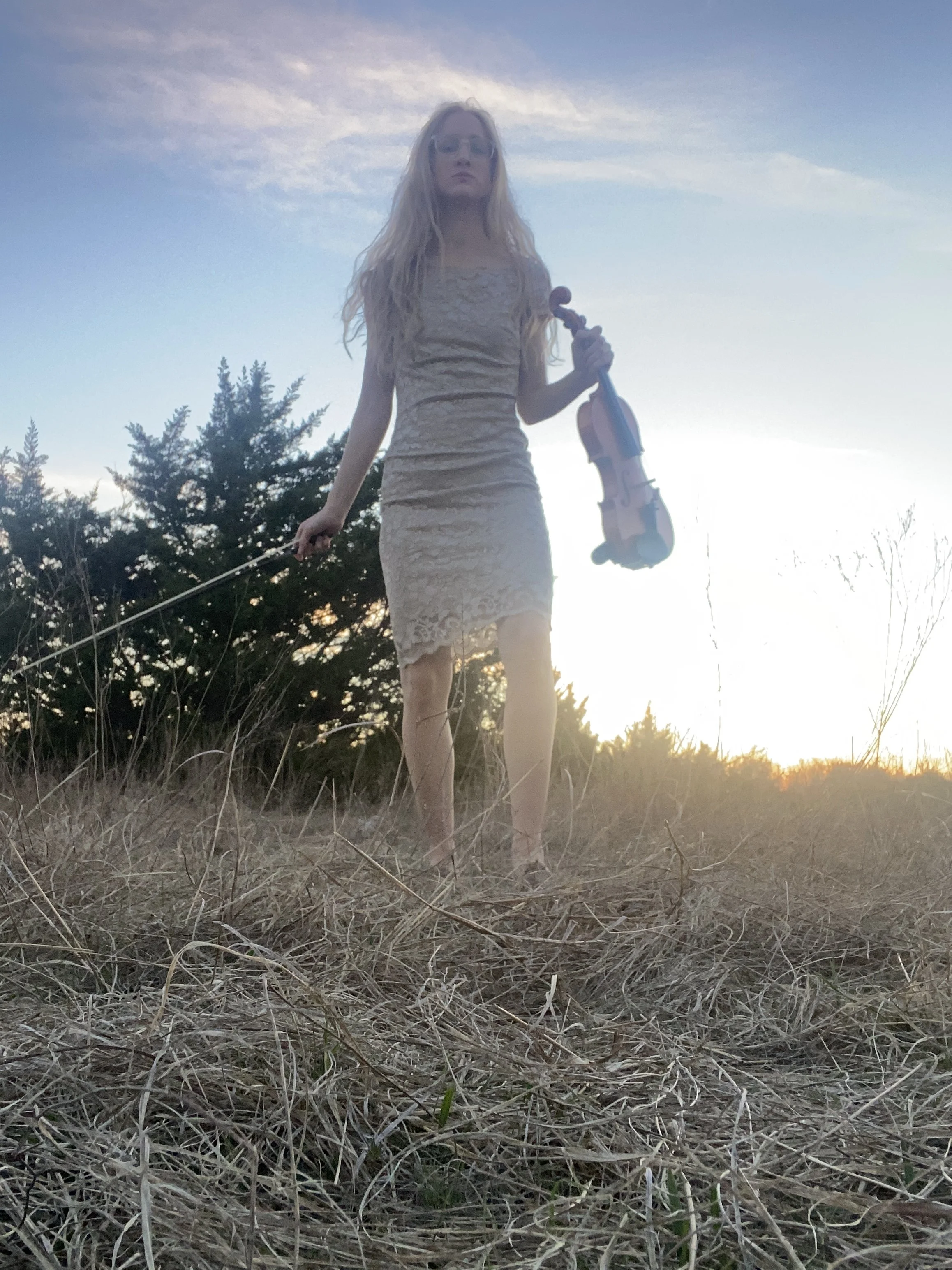 A woman with long blonde hair wearing a beige dress holding a violin in her right hand and a bow in her left, standing in a field with dry grass and trees in the background during sunset