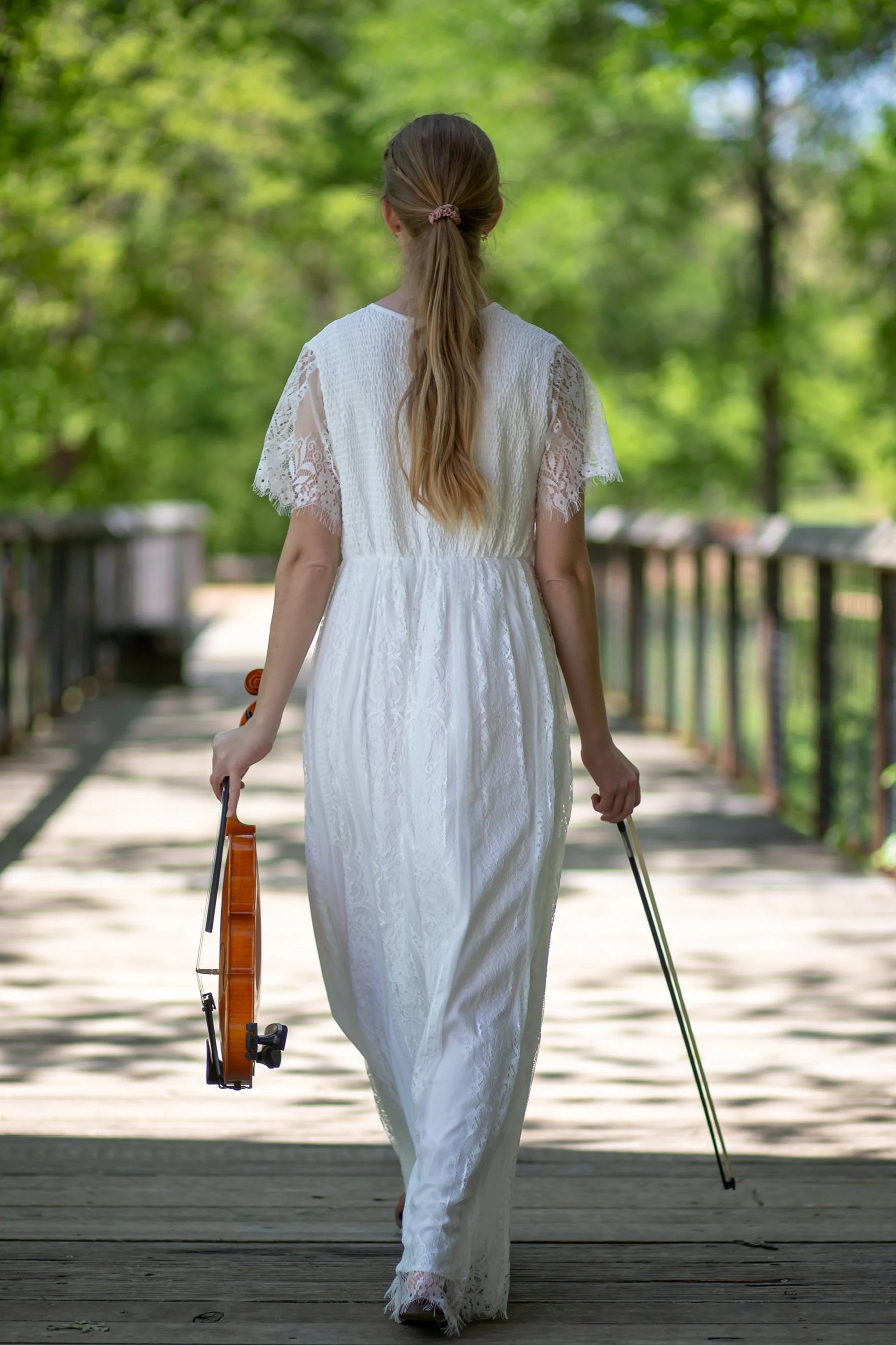 A woman in a white lace dress walking on a wooden bridge in a lush green outdoor setting, holding a violin in one hand and a bow in the other, with her back facing the camera.