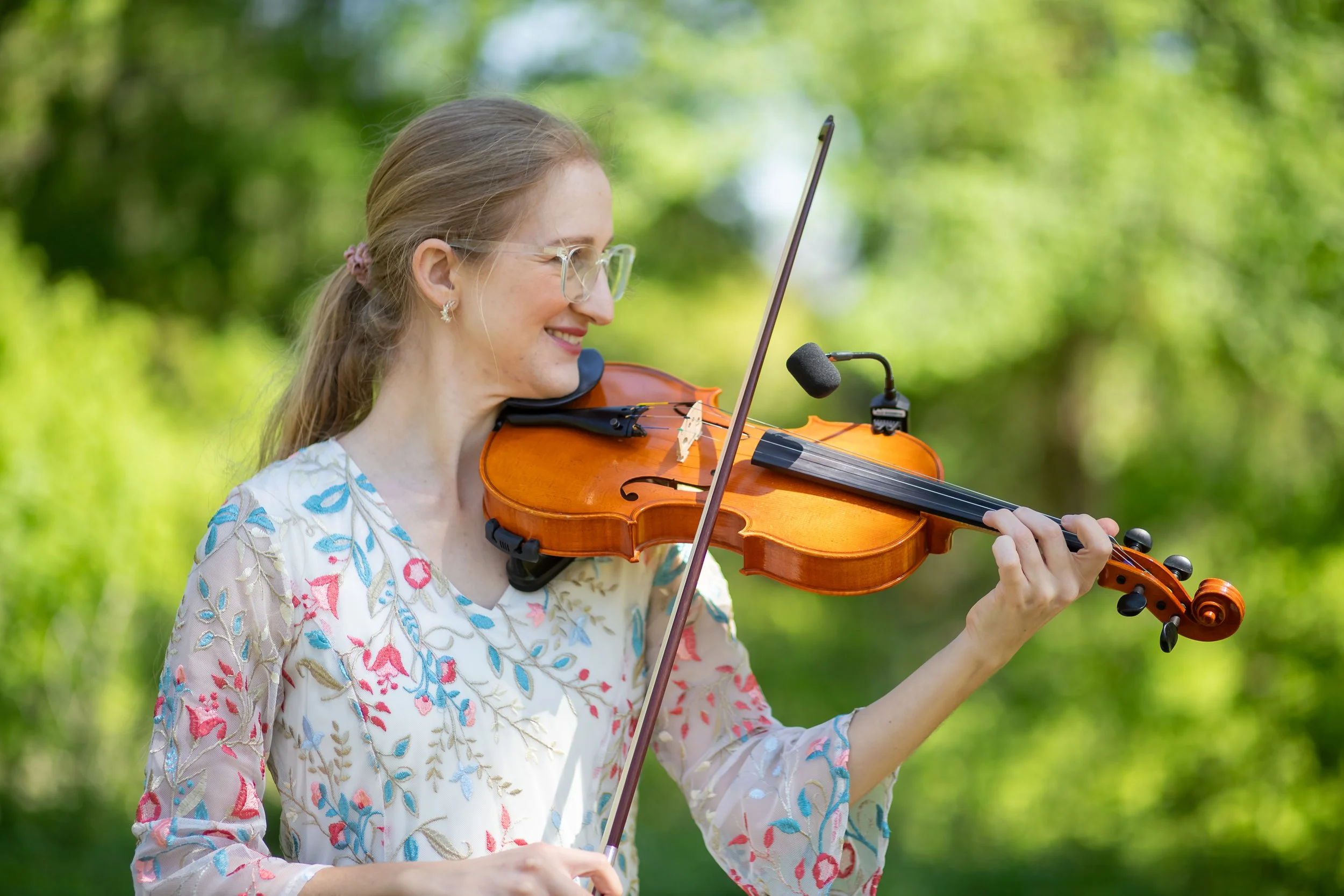 A woman with glasses playing a violin outdoors in a green, forested area.