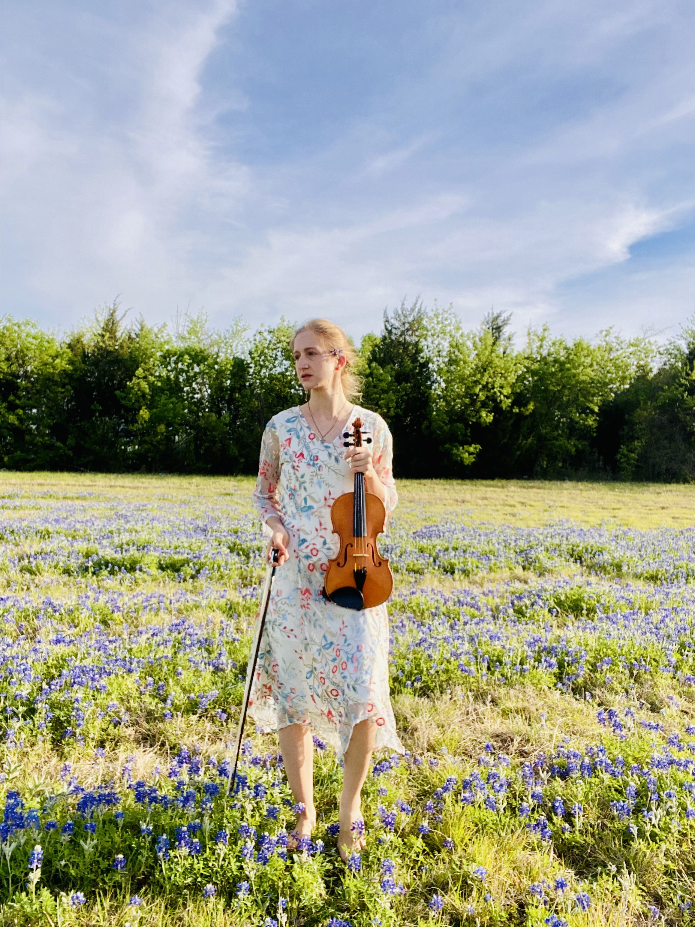 A woman standing in a field of bluebonnets, holding a violin and a bow, with a line of green trees and a blue sky with some clouds in the background.