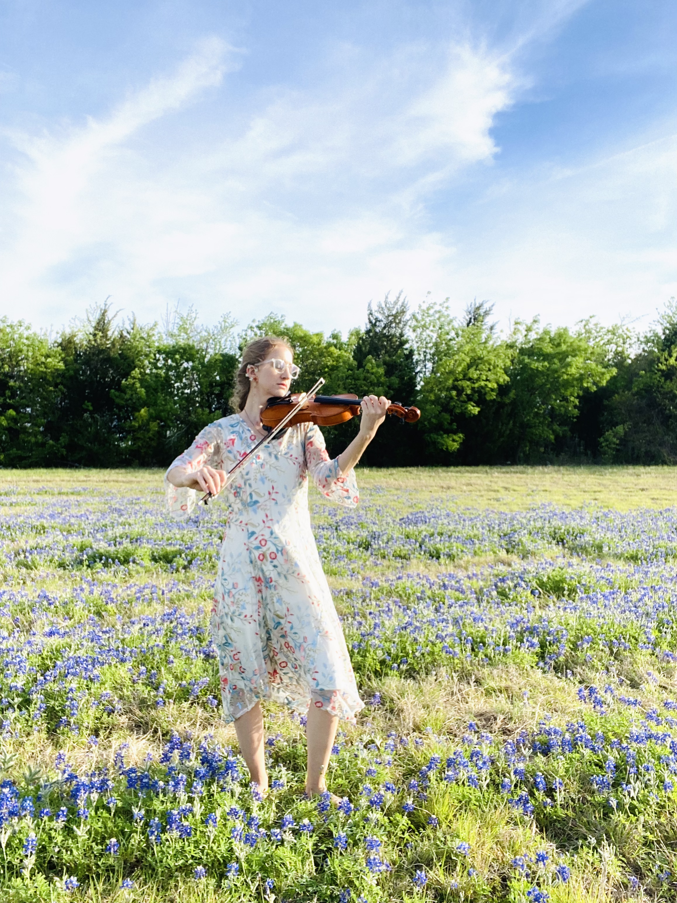 A woman in a floral dress playing the violin in a field of blue flowers, with trees and a blue sky with clouds in the background.