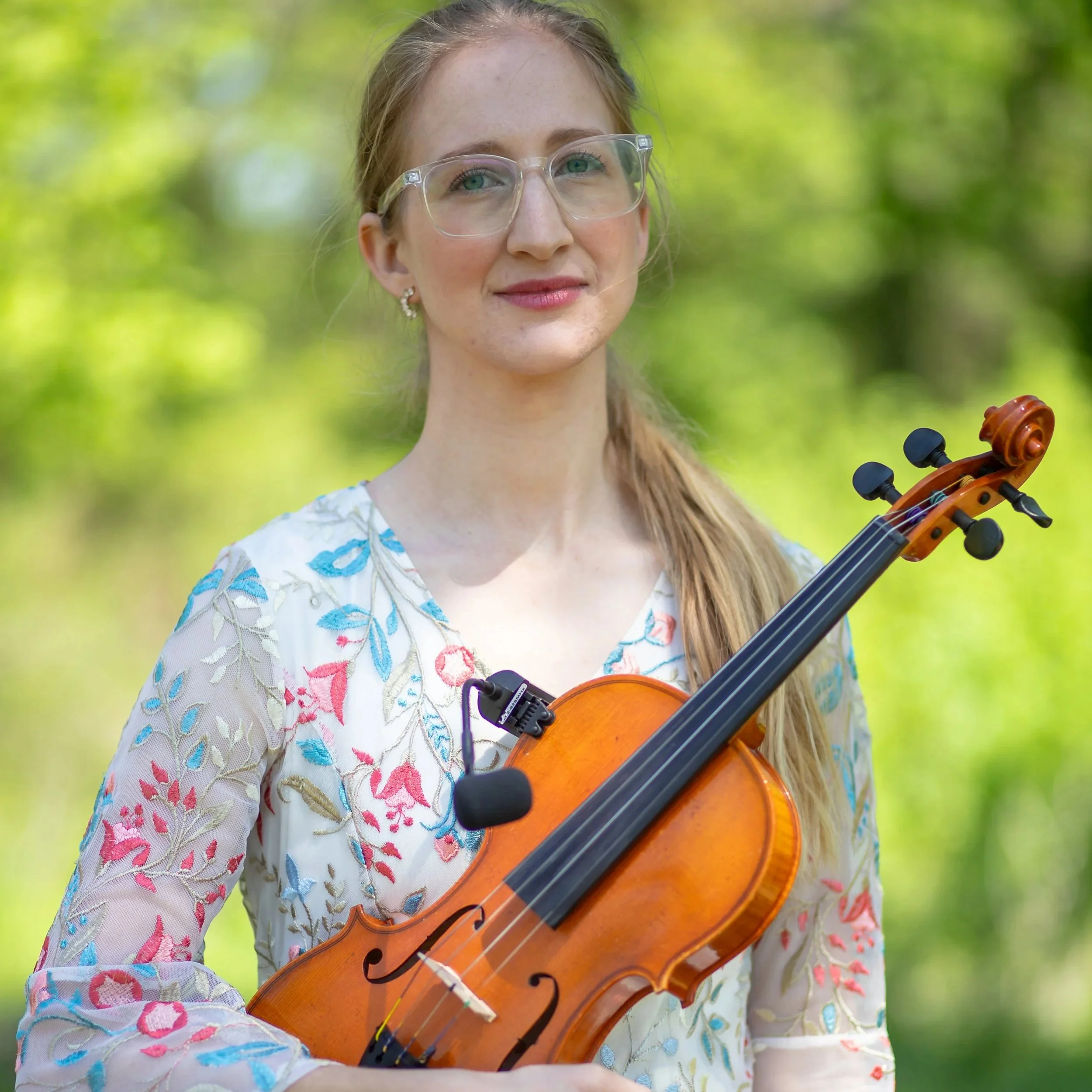 A young woman with blonde hair and glasses holding a violin outdoors with green blurred background.