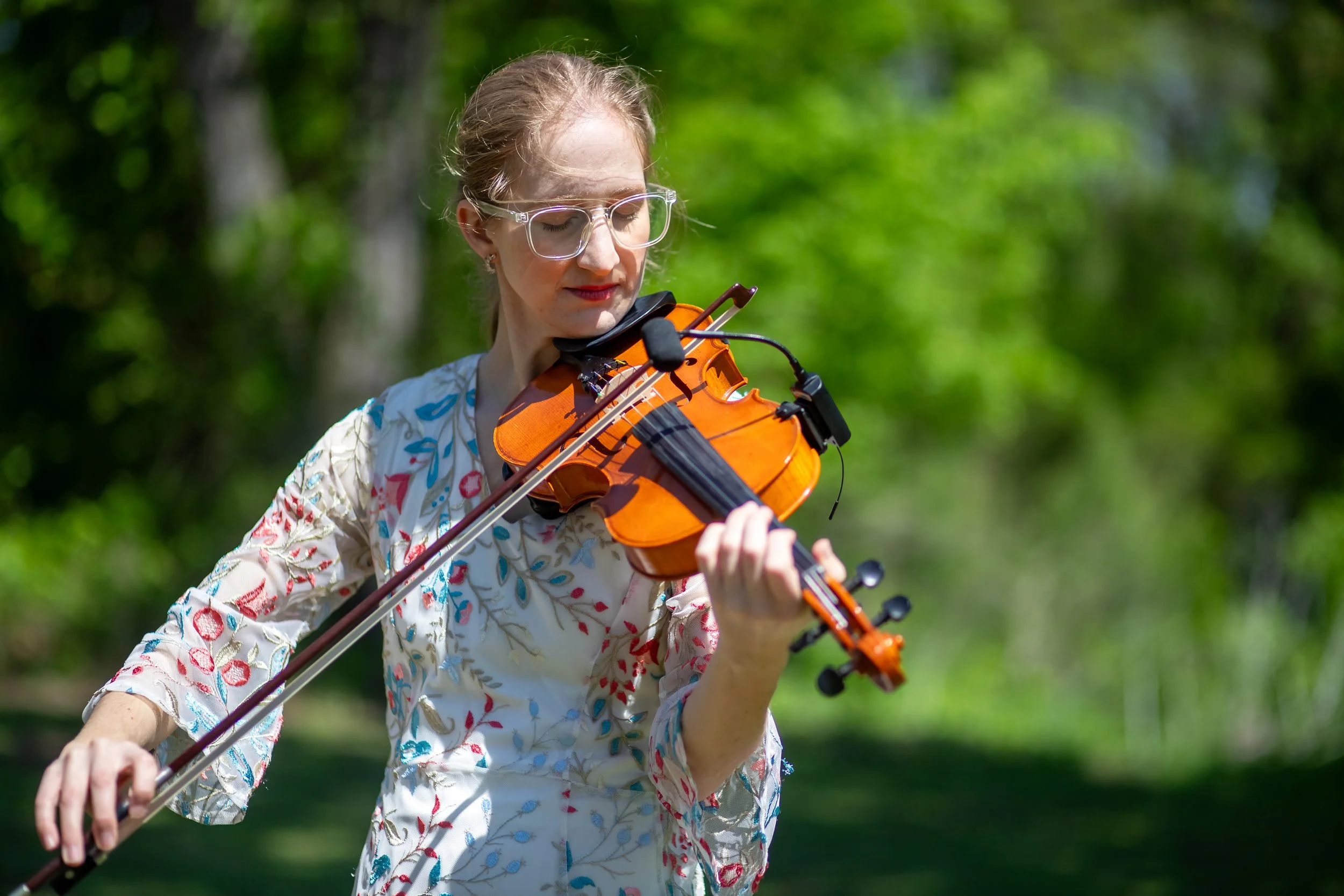 A woman with dark blonde hair, wearing glasses and a patterned floral dress, playing a violin outdoors in a green, wooded area.