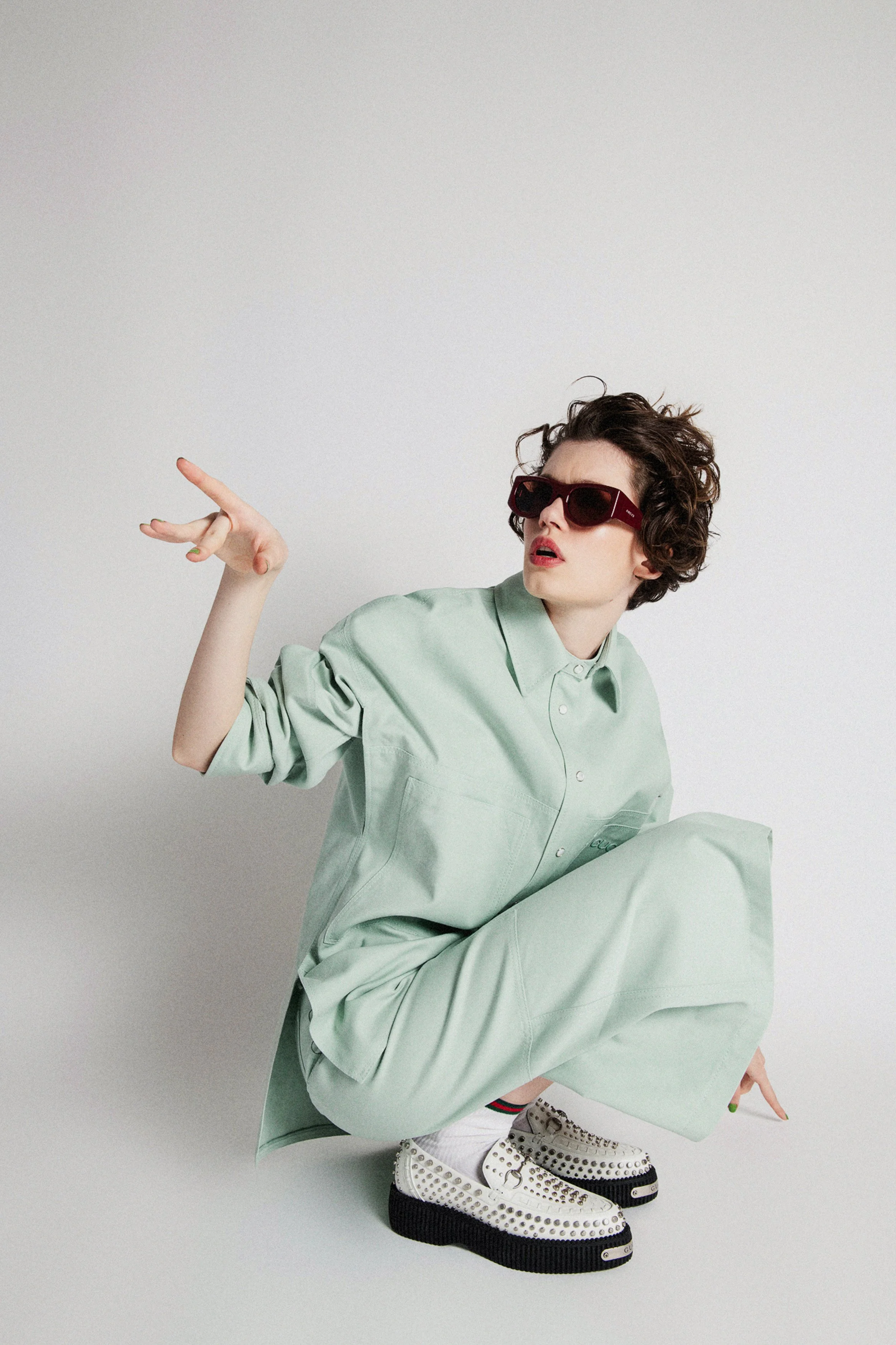 A woman with short curly hair wearing sunglasses, a light green shirt and pants, white shoes with studs, and socks, posing against a plain background.