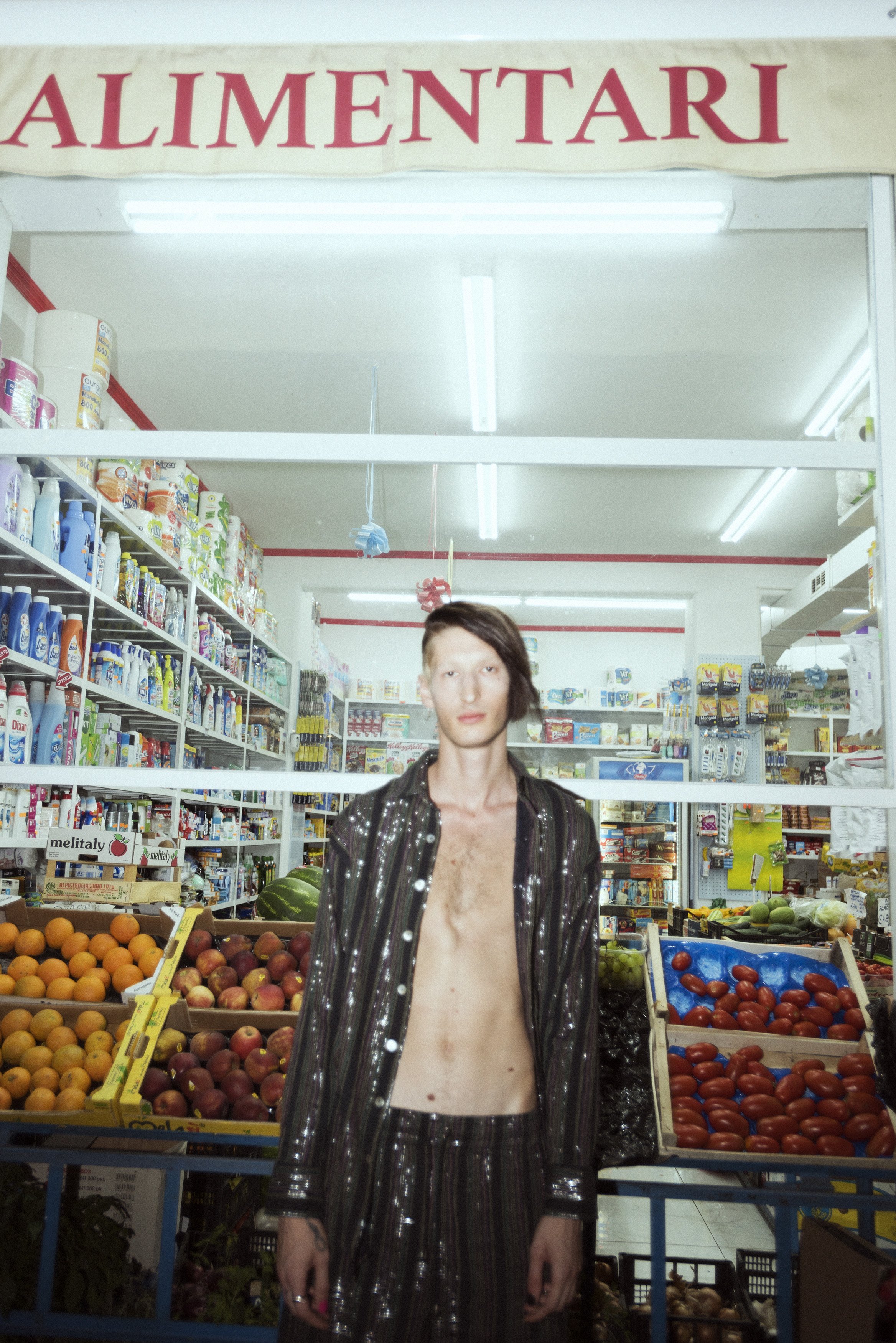 A person with a gaunt appearance, wearing a shiny, striped, partially unbuttoned jacket and matching pants, standing in front of a produce section in a grocery store with a sign reading 'ALIMENTARI' above. The store shelves behind are stocked with va
