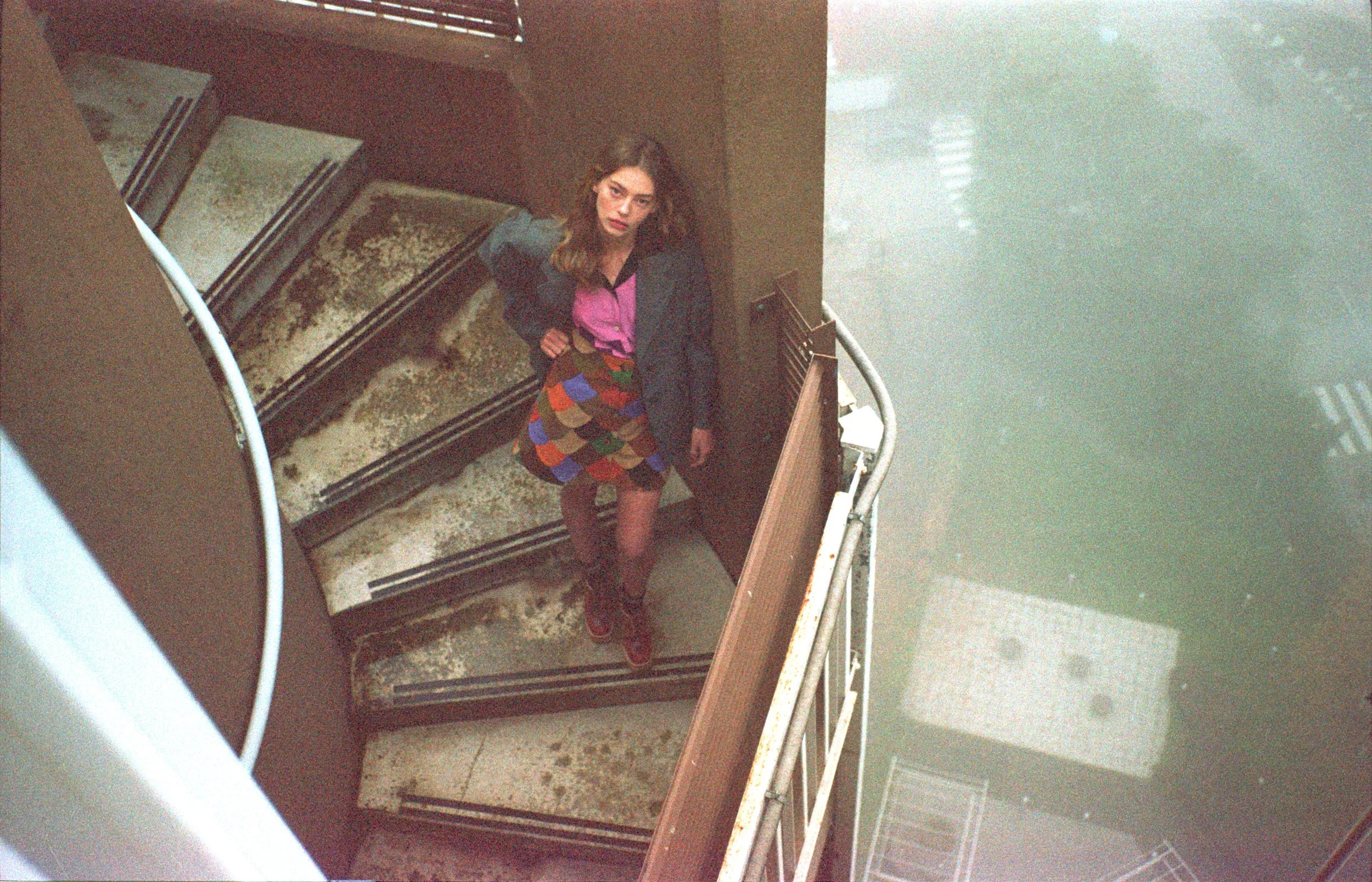 A young woman with wavy brown hair standing on a staircase looking up at the camera, wearing a colorful patterned skirt, a pink blouse, and a dark blazer.