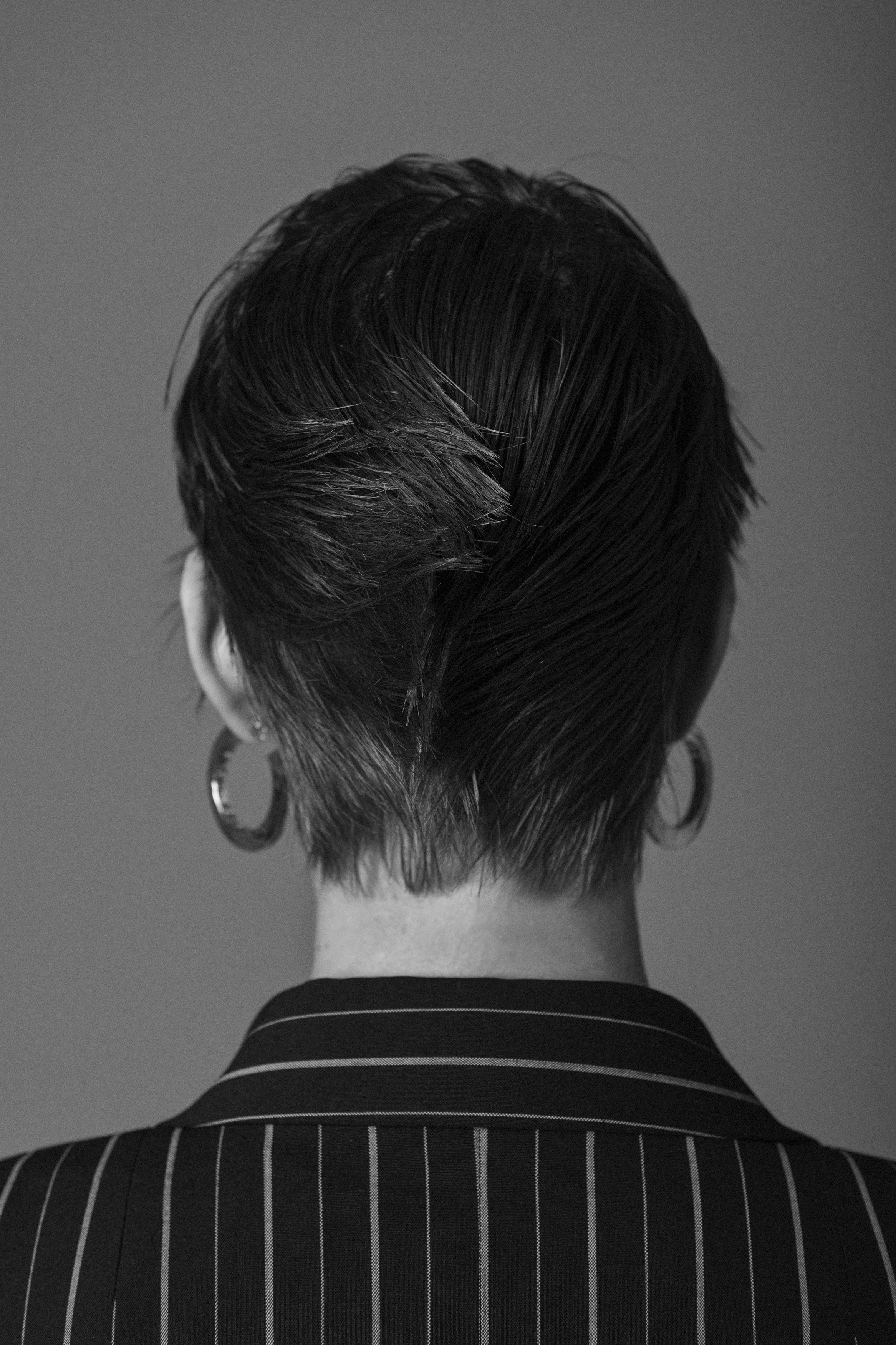 Back view of a person with short, dark, wet hair, wearing hoop earrings and a pinstripe blazer.