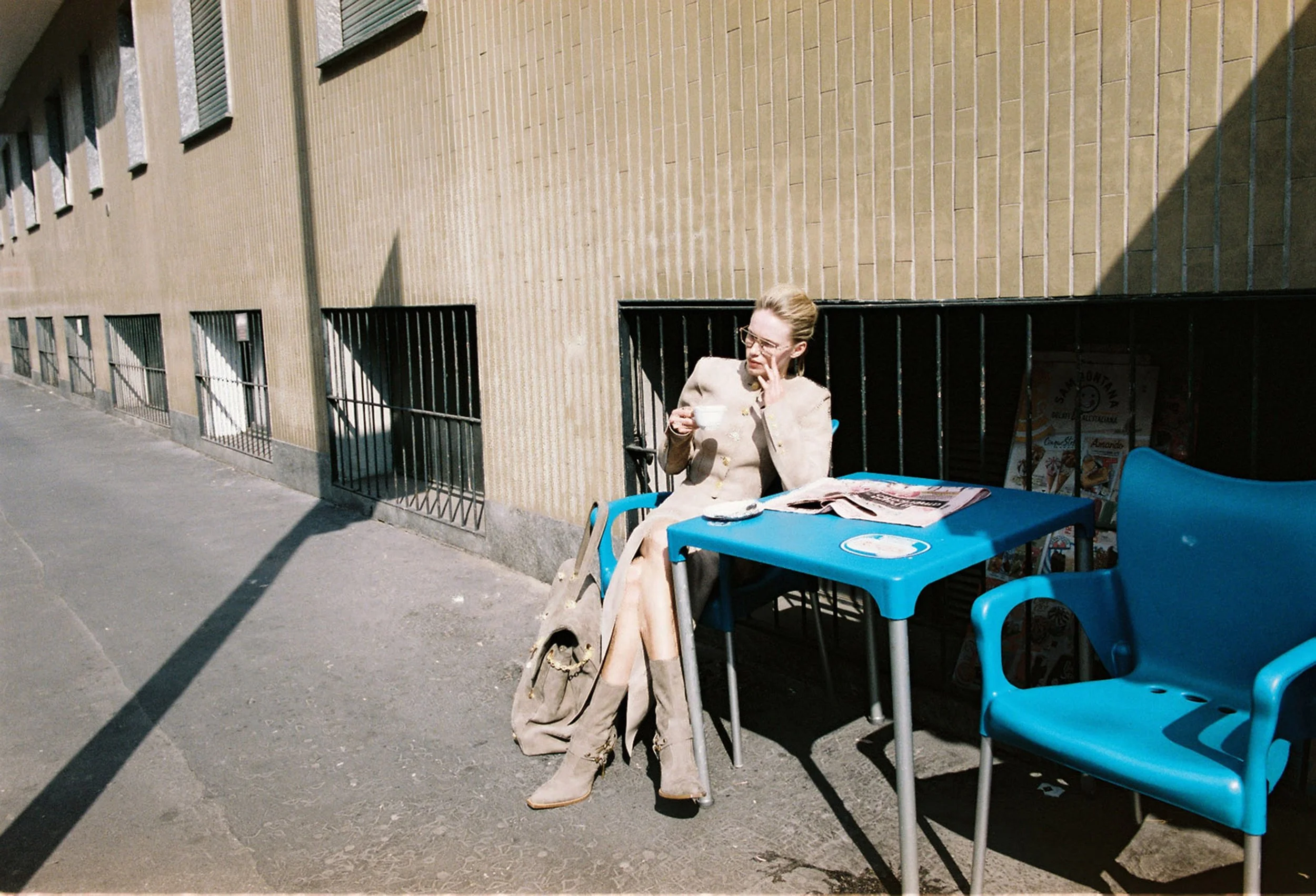 A woman sitting outside at a blue table, drinking from a cup, with a newspaper on the table. She is wearing a beige coat and high boots. There are blue chairs and a backpack beside her. The scene is on a sidewalk next to a brick building with barred 