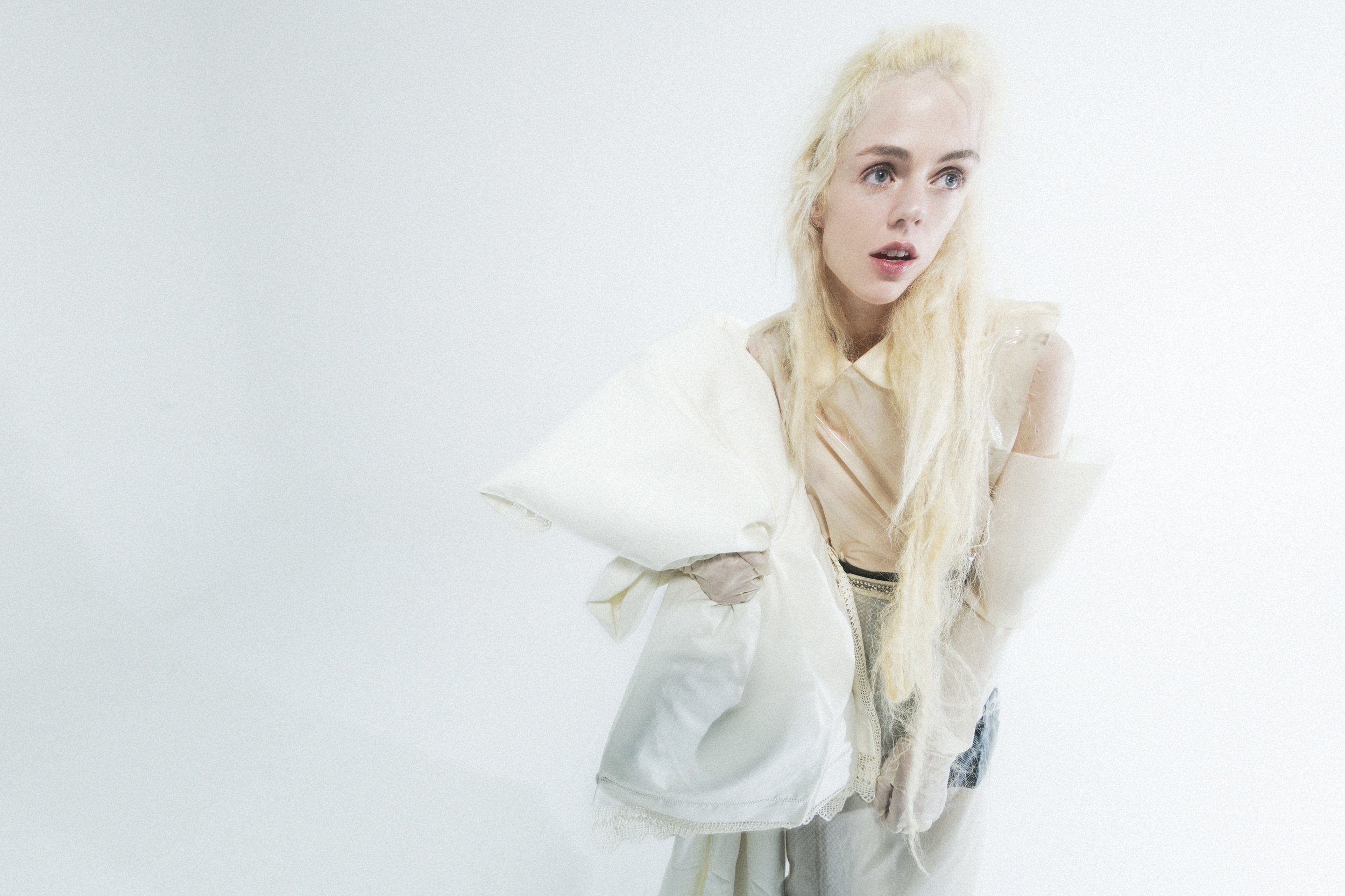 A blonde woman with long, wavy hair looking surprised or confused, dressed in pastel or light-colored clothing, holding a white garment or bag, standing against a plain white background.