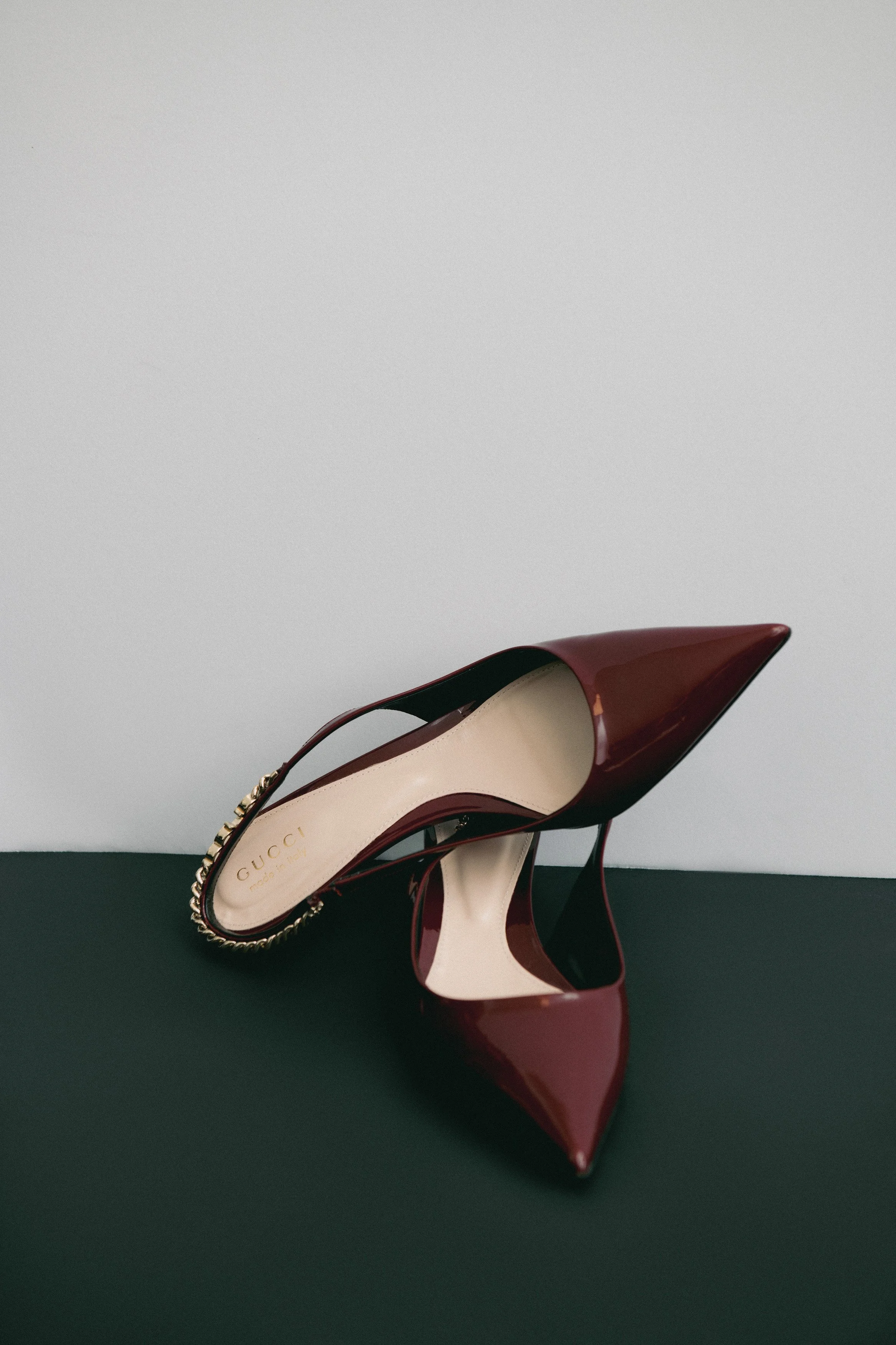 A  Gucci pair of red patent leather high heels with a pointed toe, one shoe resting on its side and the other upright, against a white and dark green background.