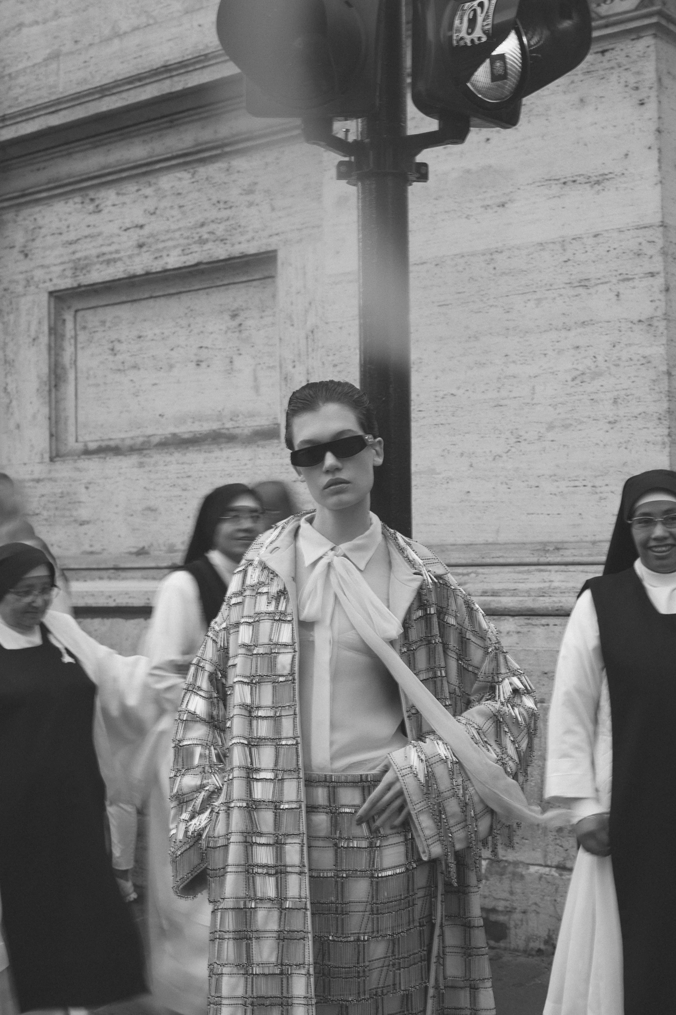 A woman dressed in a plaid patterned outfit and sunglasses standing among nuns on a city street in front of a building wall and traffic light.