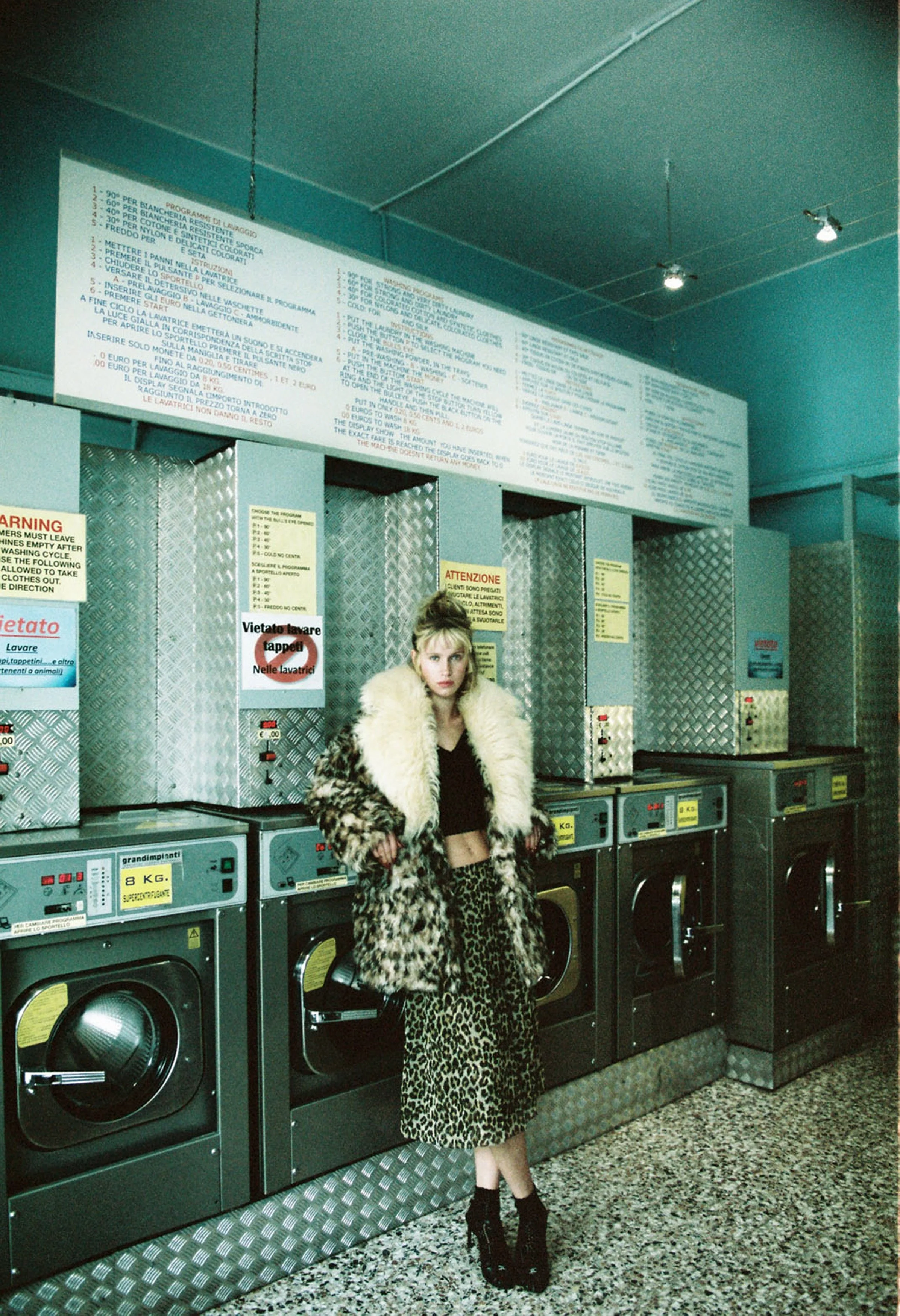 A young woman in leopard print pants, black top, and furry coat standing in front of coin-operated laundry machines in a laundromat.