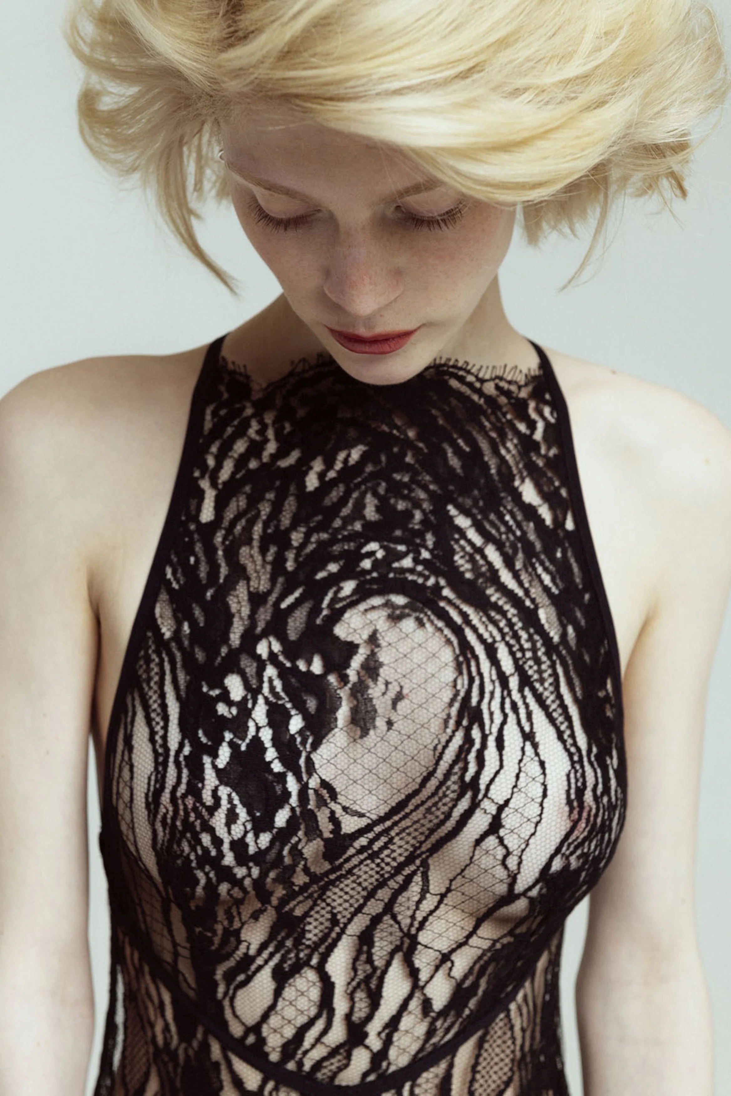 A woman with short blonde hair looking downward, wearing a black lace top with intricate patterns.