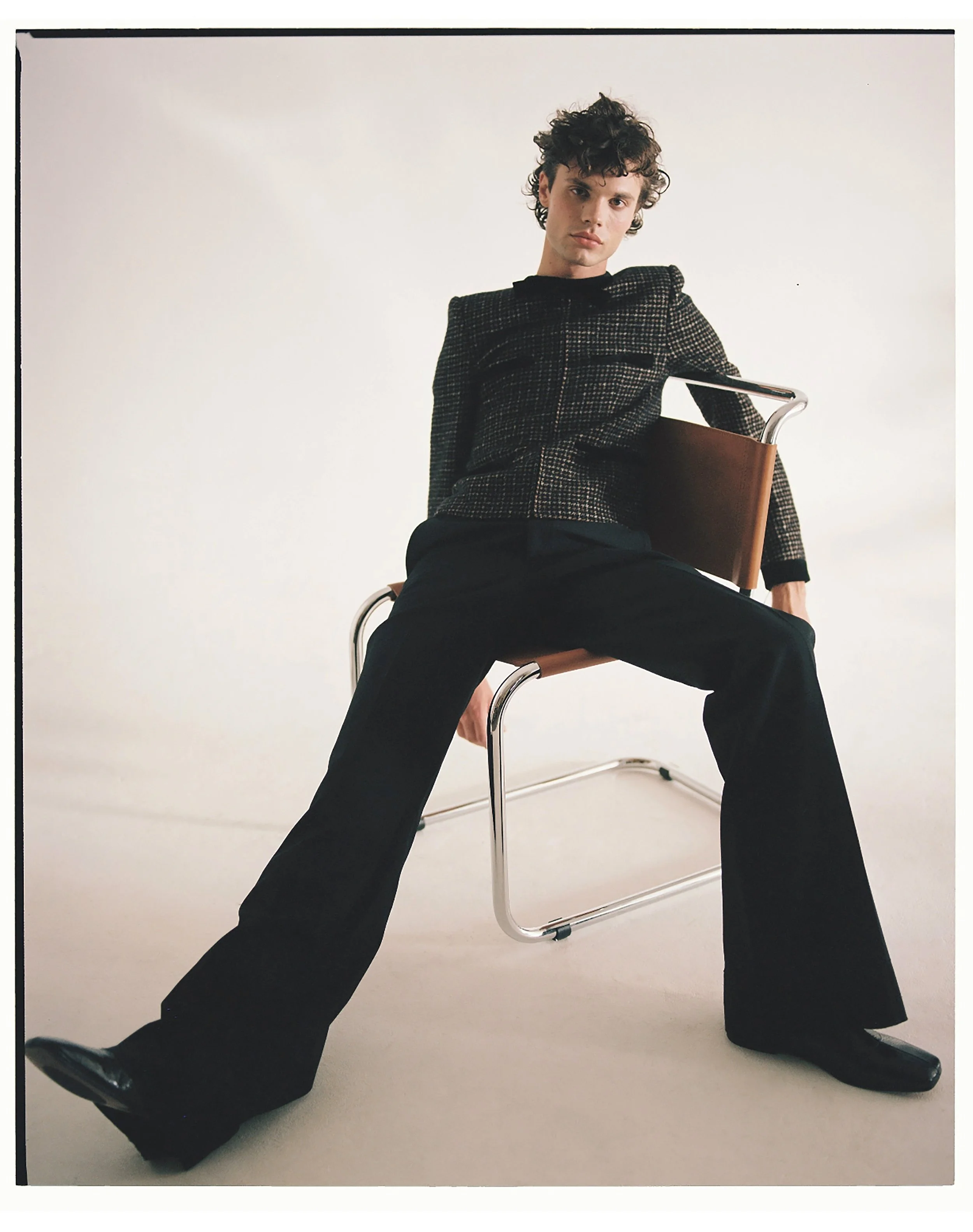 Young man with curly hair seated on a brown chair with steel armrests, wearing a black and brown patterned blazer and black wide-leg pants, against a plain white background.