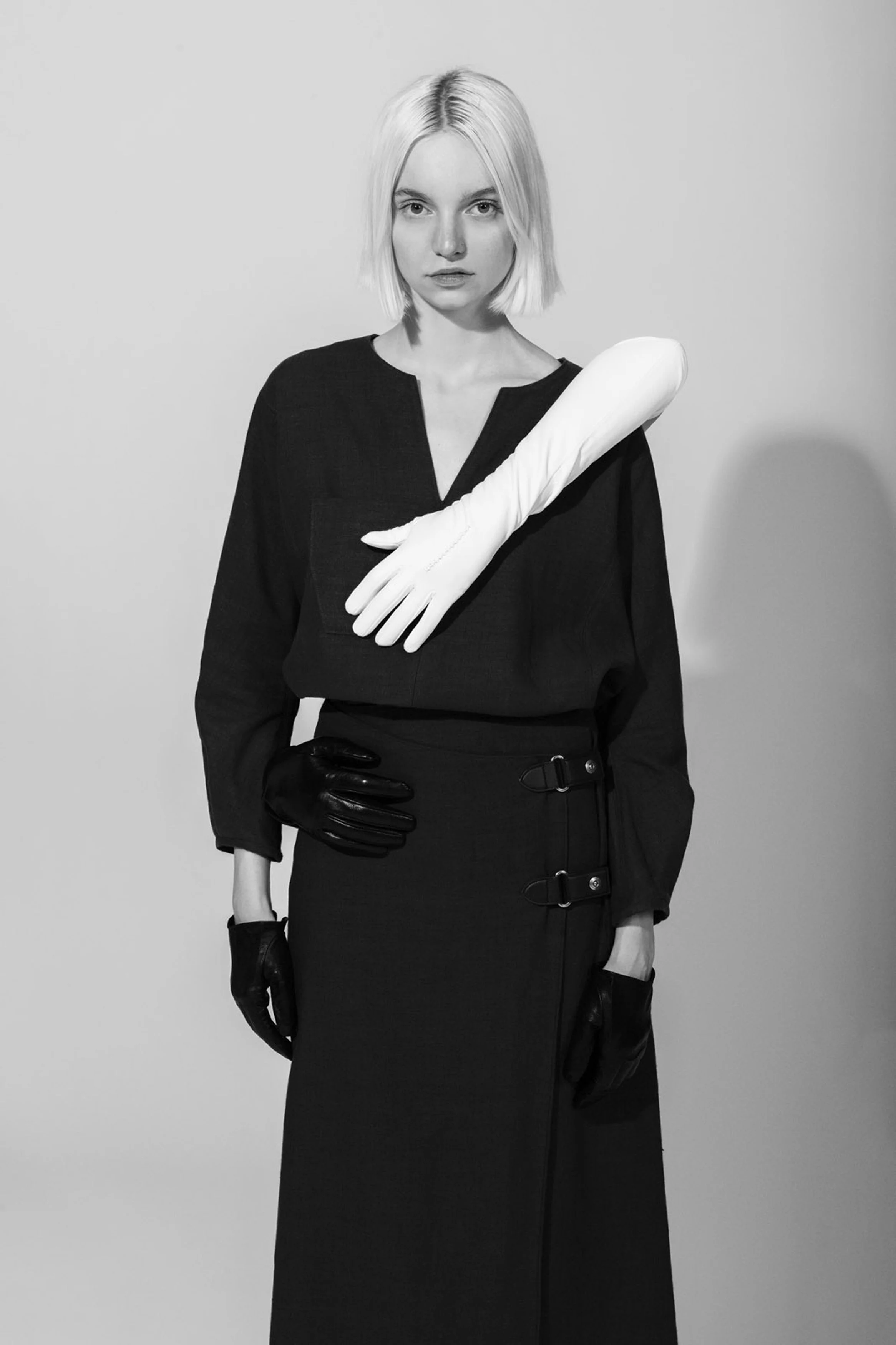 Black and white photo of a woman with short blonde hair, wearing a long dark dress, black gloves, and a white glove on one arm, standing against a plain background.