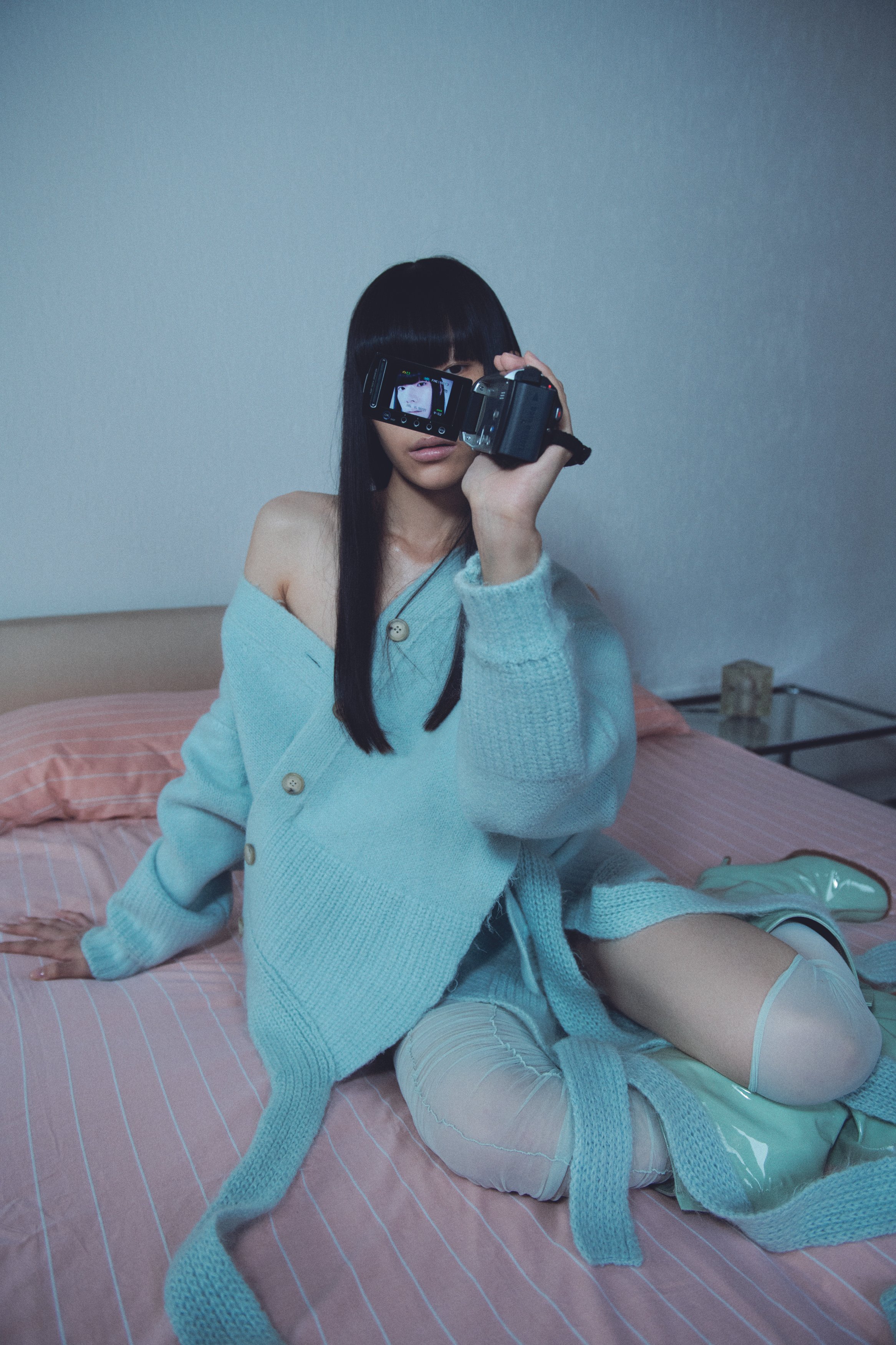 A woman with long black hair and bangs sitting on a pink bed with striped sheets, holding a camera up to her face. She is wearing a light blue oversized sweater, ripped jeans, and shiny knee-high boots.