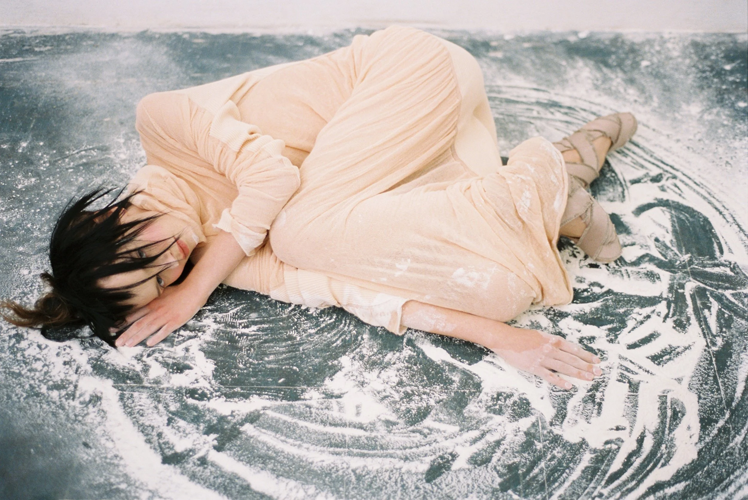 A woman lying on a black surface covered in white powder, wearing a light pink dress and ballet shoes, with dark hair and an expressive pose.