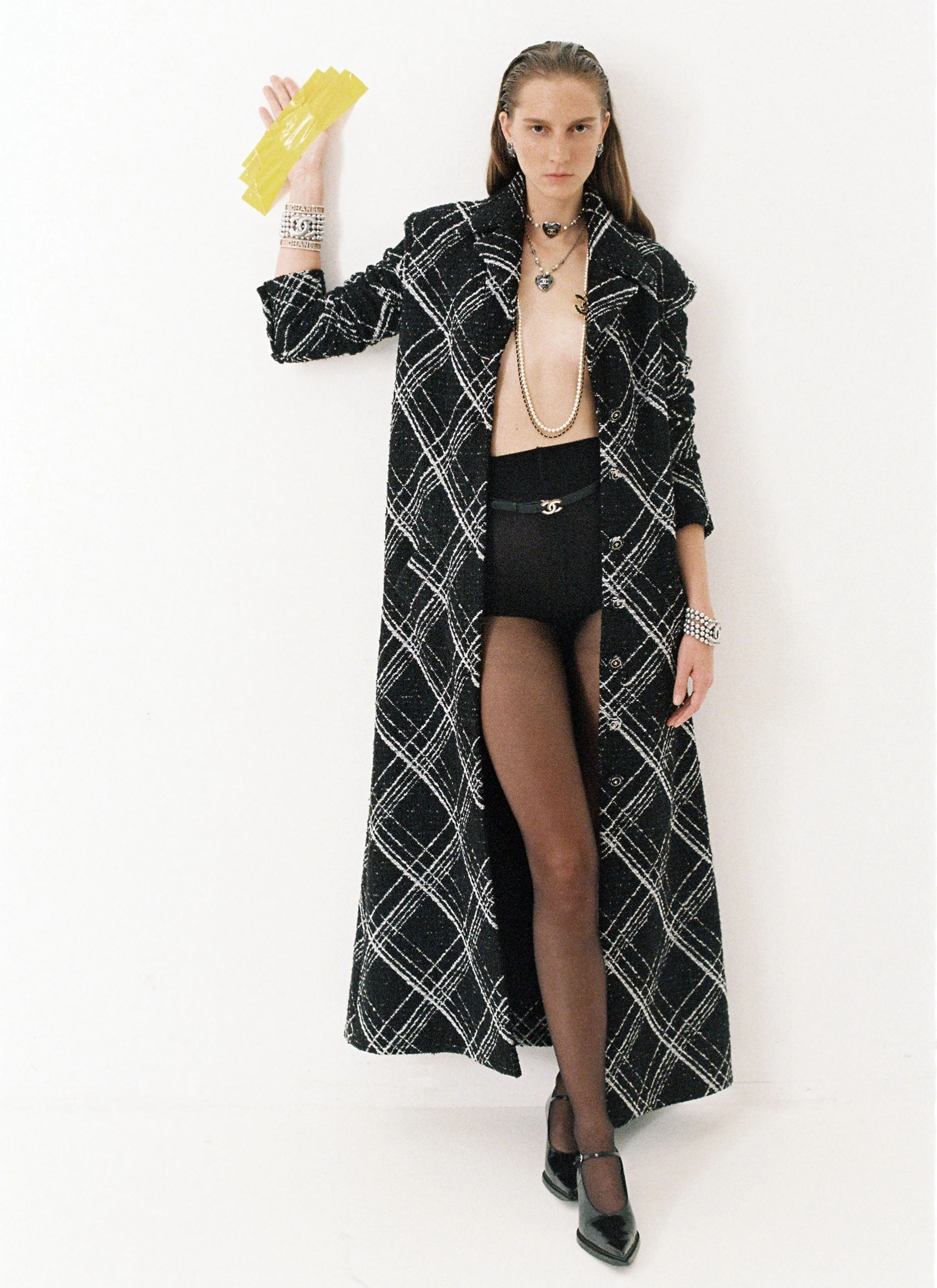 Fashion model in a long black and white plaid coat, black shorts, sheer black tights, and black high heels, holding yellow paper fans, against a white background.