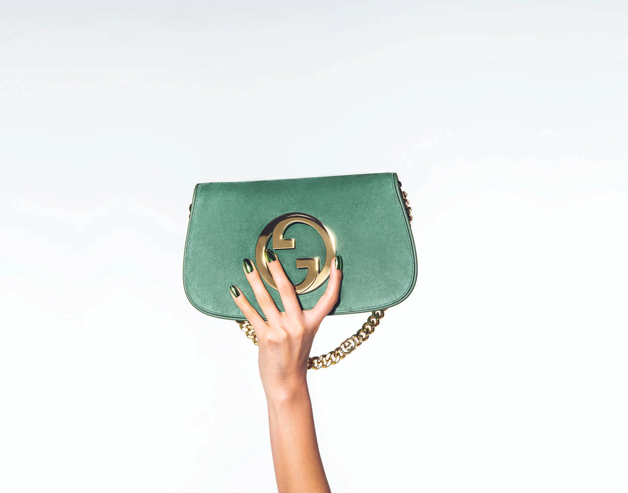 A person with manicured nails holding a green Gucci handbag with a gold logo against a plain, light background.
