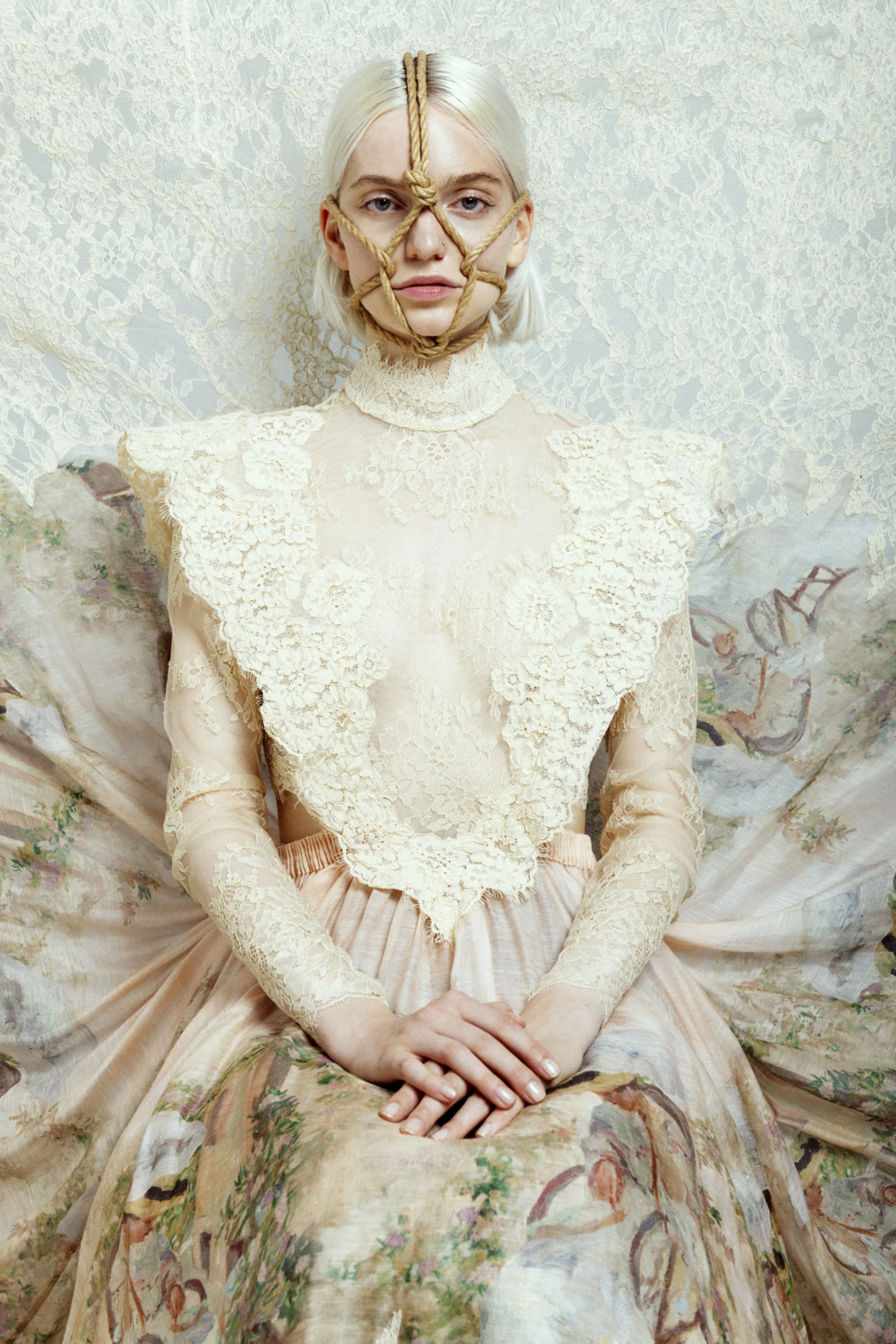 A woman with platinum blonde hair wears a lace top and a floral patterned skirt, sitting with her hands folded on her lap, and a rope tied around her face in a star-shaped pattern, against a lace background.