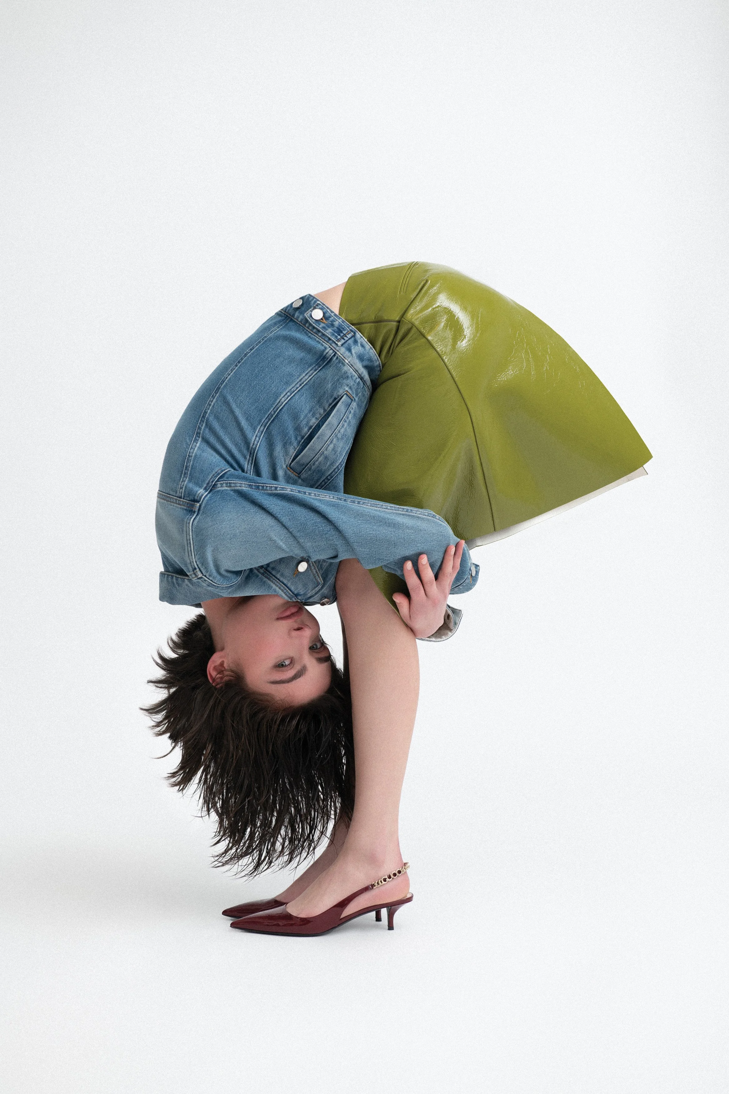 A person with short dark hair bending forward, wearing a denim shirt and red high heels, leaning over a green leather chair in an unusual upside-down pose.